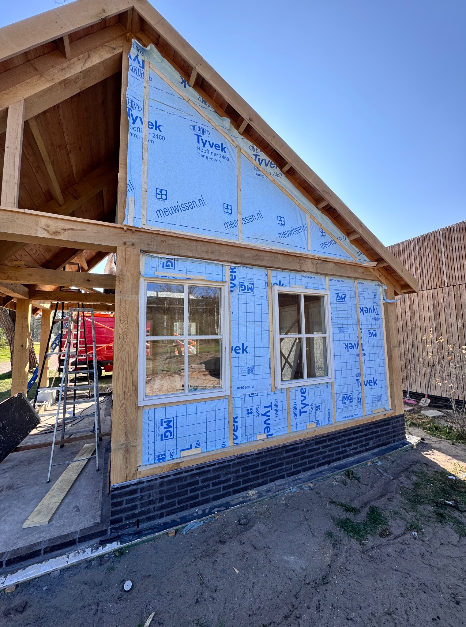 Konstrukcja szkieletowa budynku w trakcie budowy. Widoczne drewniane belki, okna, folia izolacyjna Tyvek i fragment muru z ciemnej cegły. Na pierwszym planie ziemia.