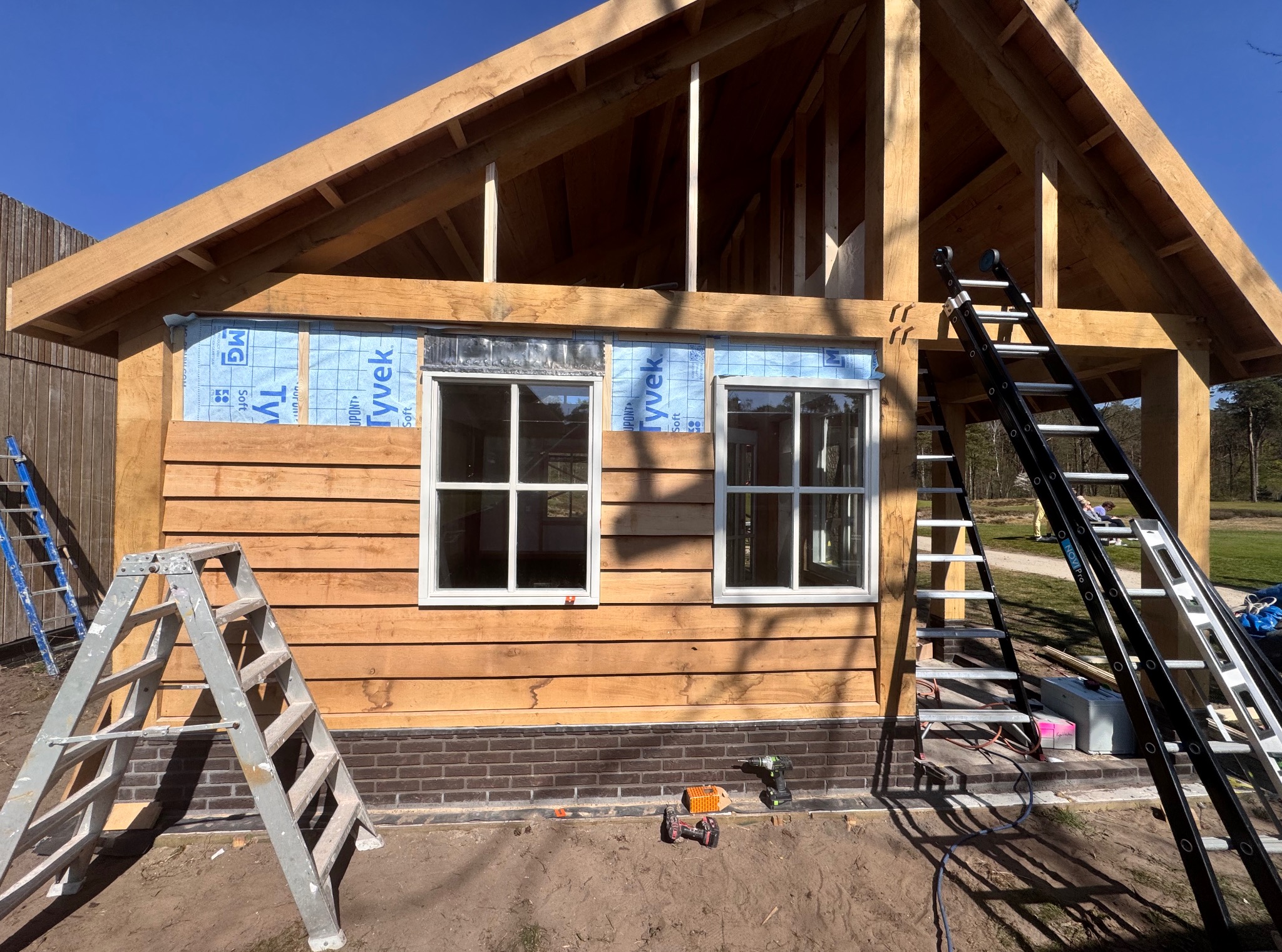 Surowy szkielet drewnianej konstrukcji z oknami, zabezpieczony folią, z dwiema drabinami i narzędziami, ukazujący etap budowy na zewnątrz.