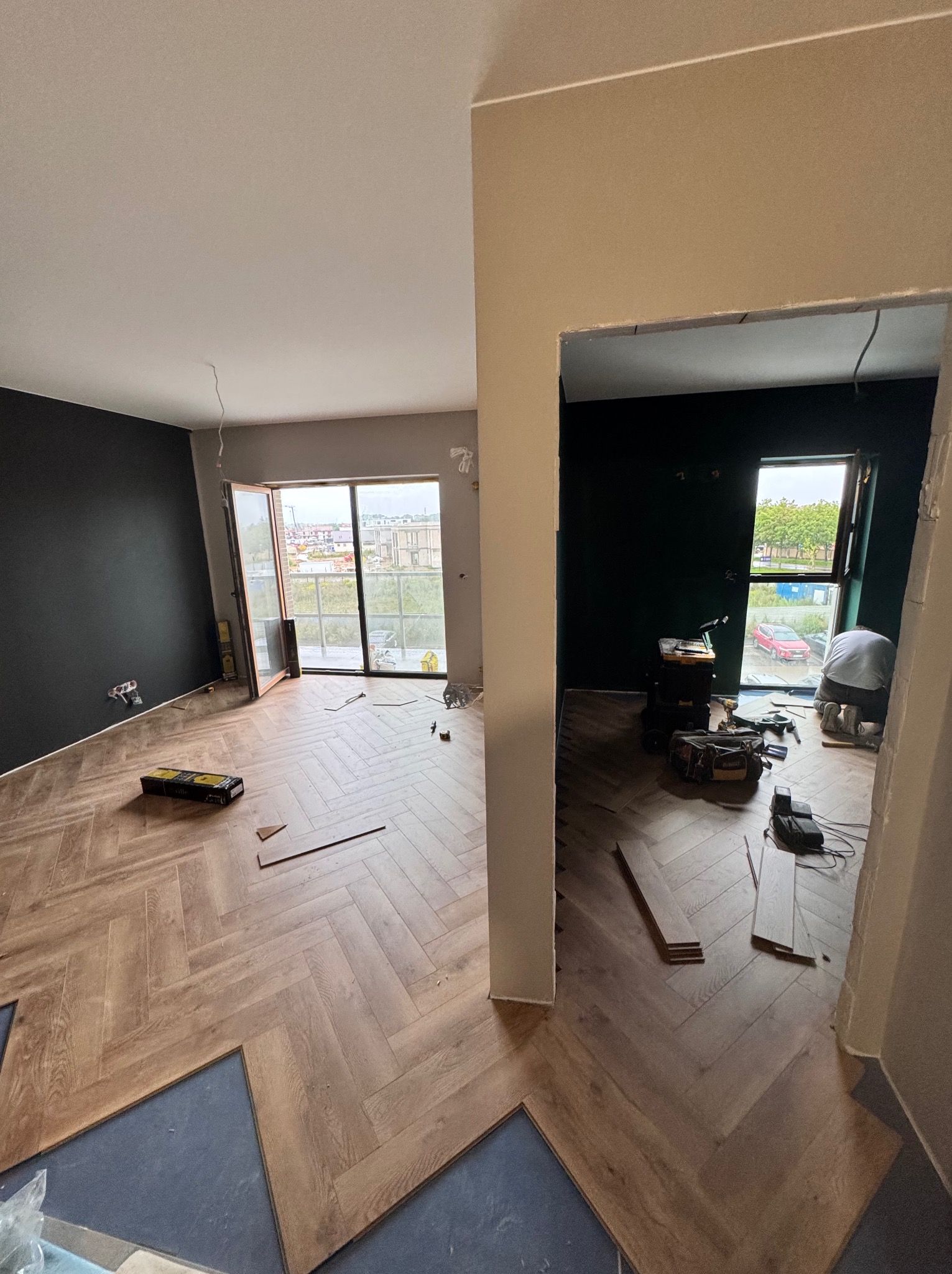 Układanie parkietu w jodełkę francuską w mieszkaniu w Poznaniu. Widoczne narzędzia i częściowo ułożona podłoga. Ściany w kolorach szarości i zieleni.