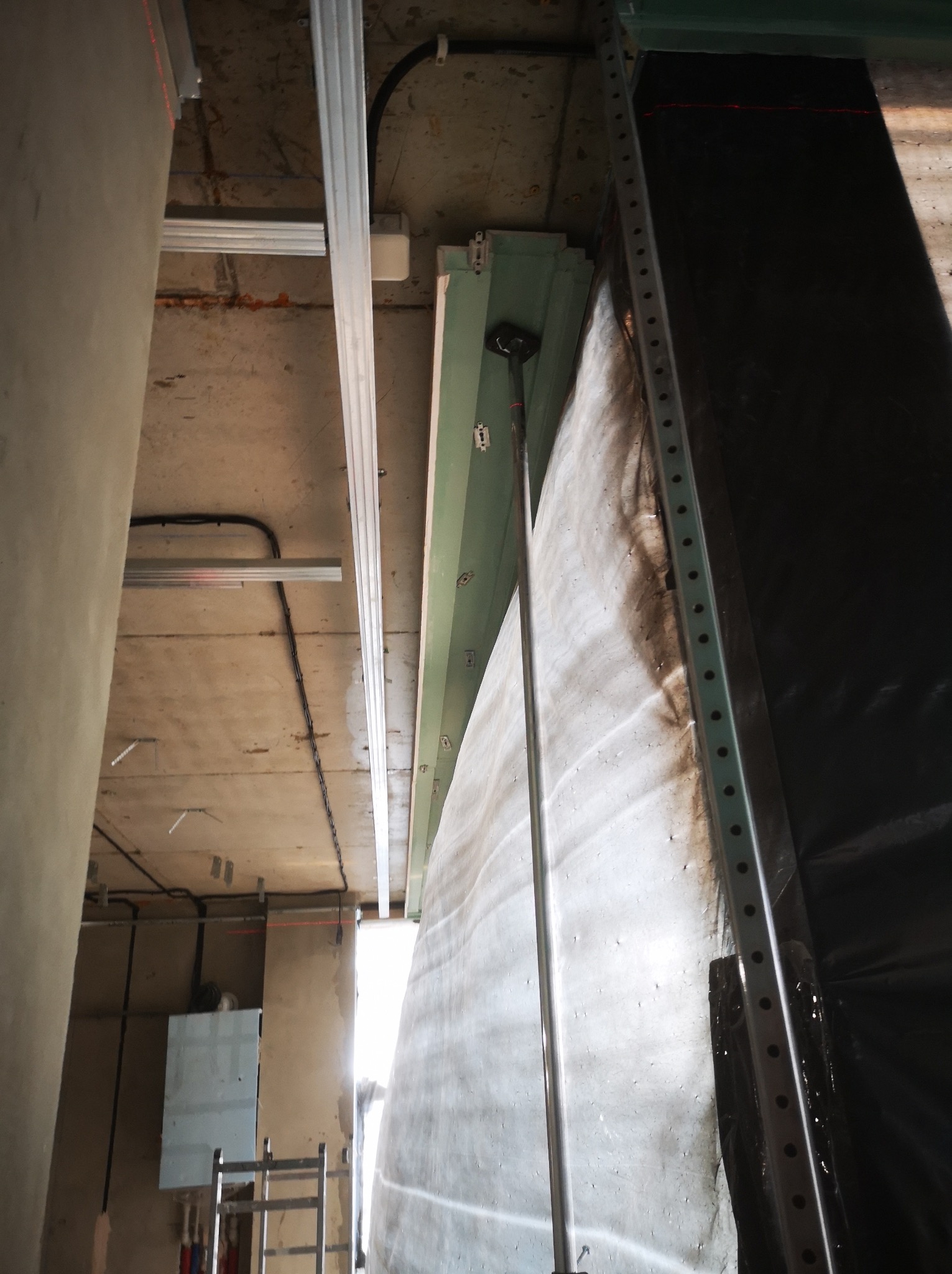 Montaż stelaża pod płytę gipsową, widoczne profile metalowe, sufit z instalacjami, ściana zabezpieczona folią. Prace wykończeniowe w surowym wnętrzu.