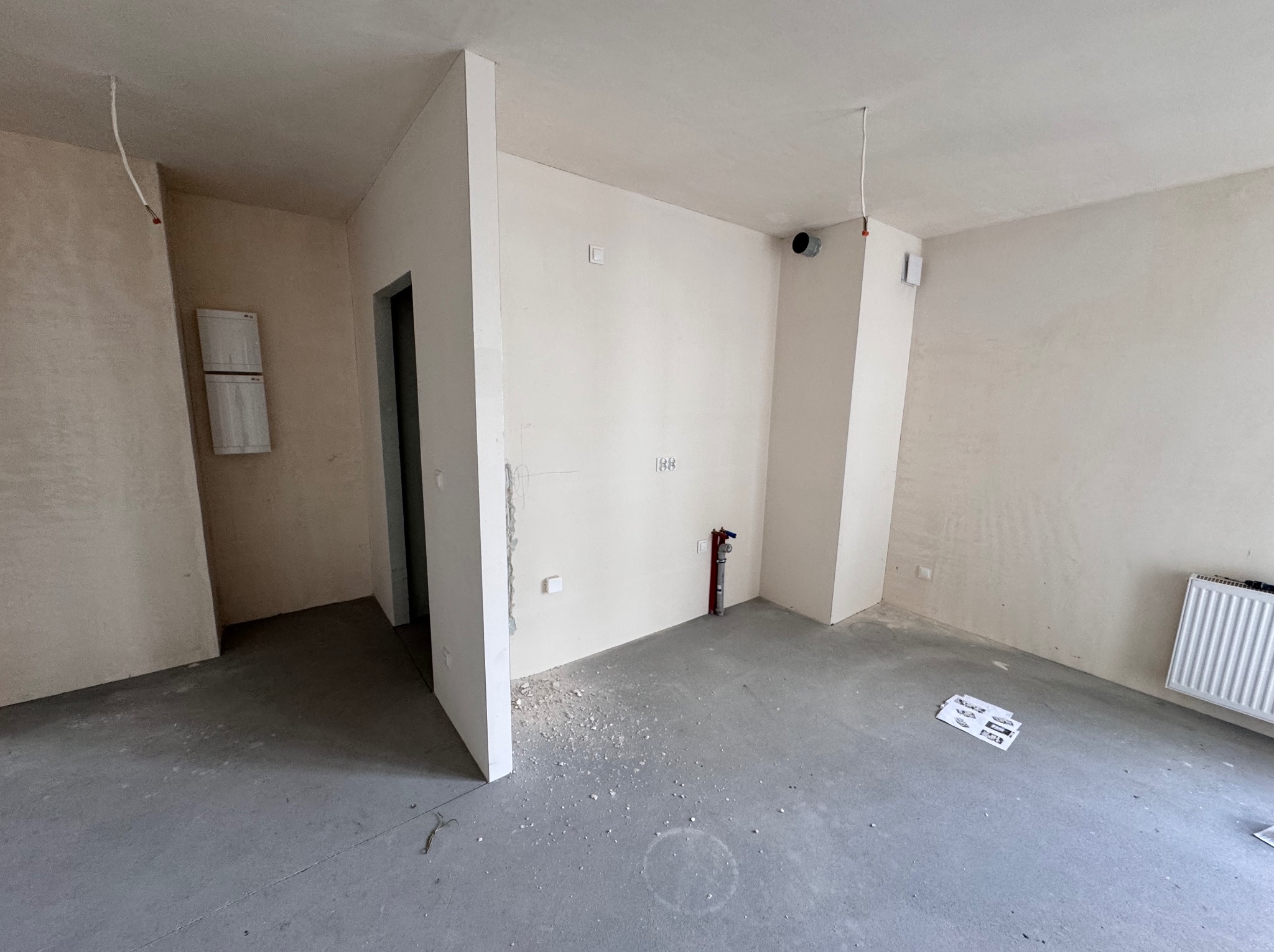 Surowe wnętrze mieszkania w trakcie remontu: gładkie ściany, betonowa podłoga, wystające kable, grzejnik i elementy instalacji. Widoczne ślady prac budowlanych.