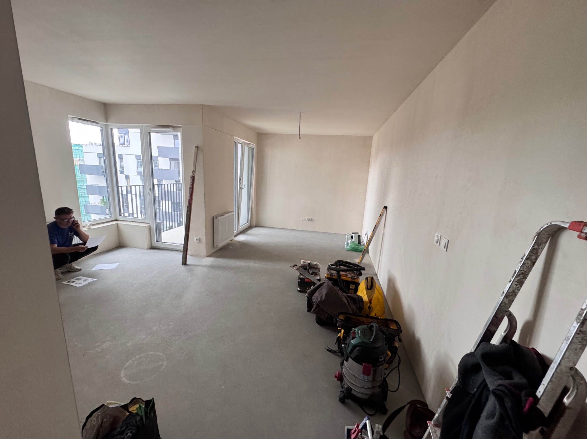 Wnętrze w trakcie prac wykończeniowych: gładkie ściany, surowa podłoga, narzędzia i drabina. Mężczyzna analizuje dokumenty przy oknie balkonowym.