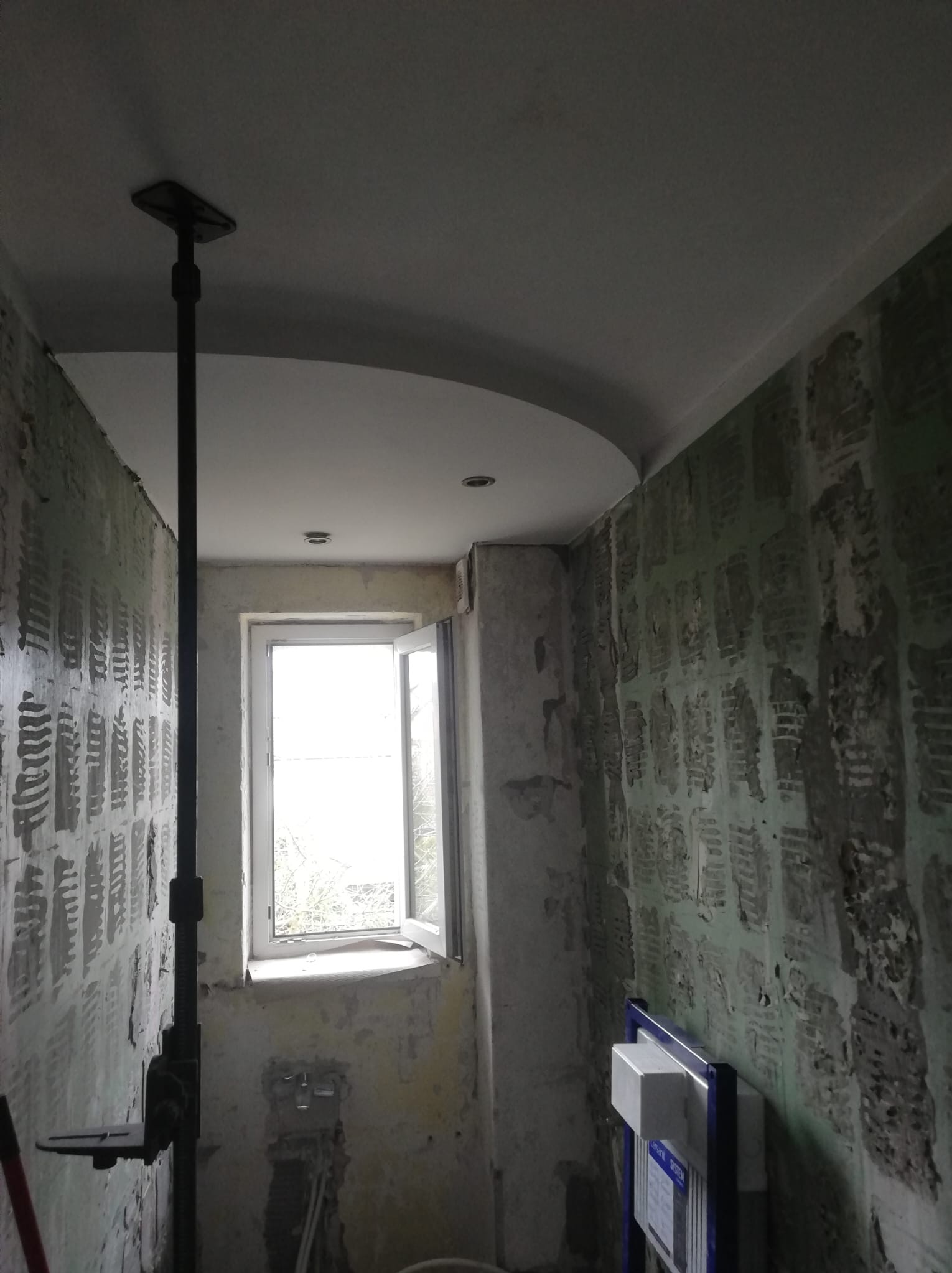 Łazienka w trakcie remontu: ściany przygotowane pod płytki, widoczne instalacje, okno. Sufit z podwieszanym elementem. Czarny statyw w rogu pomieszczenia.