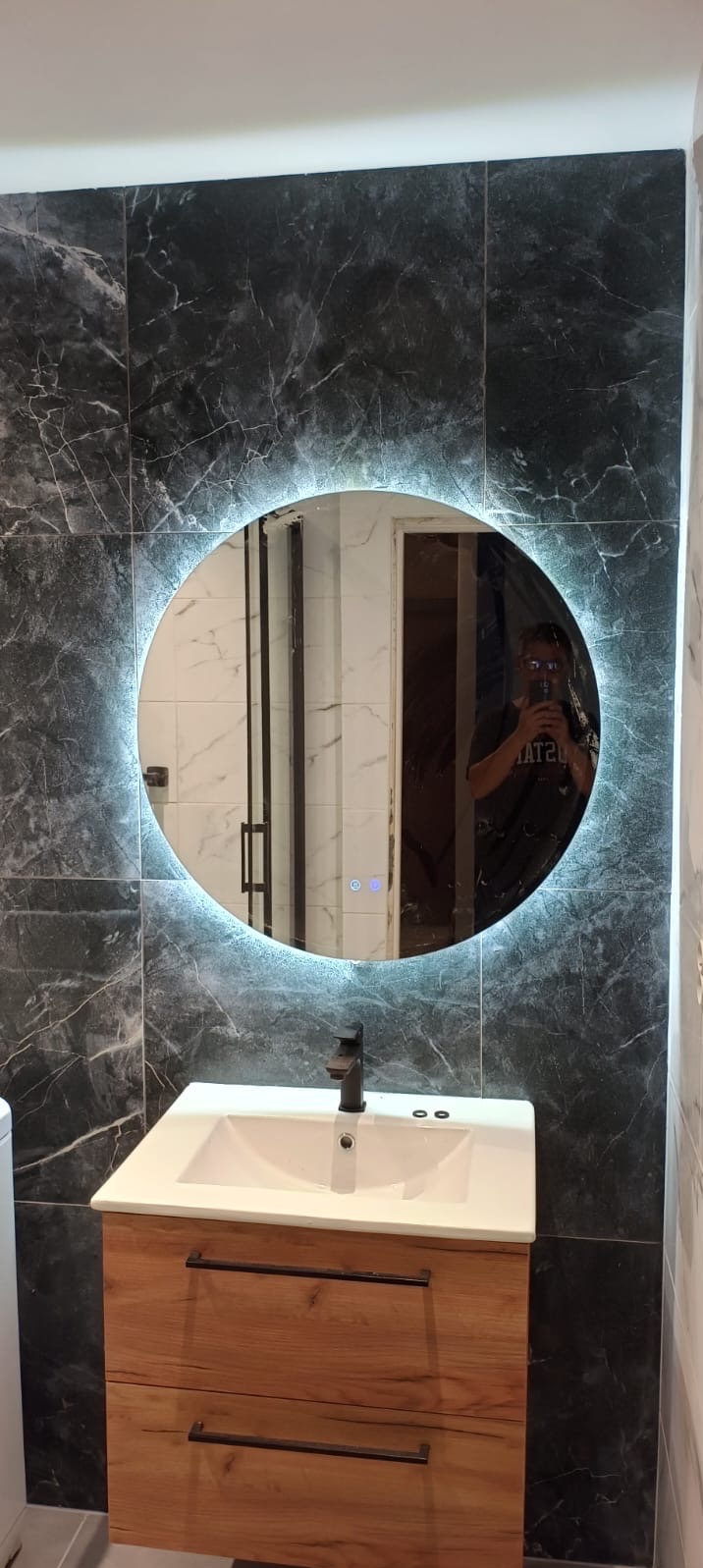 Nowoczesna łazienka: ciemne marmurowe płytki, okrągłe lustro LED, dębowa szafka z umywalką nablatową i czarna bateria. Widoczne odbicie osoby robiącej zdjęcie.