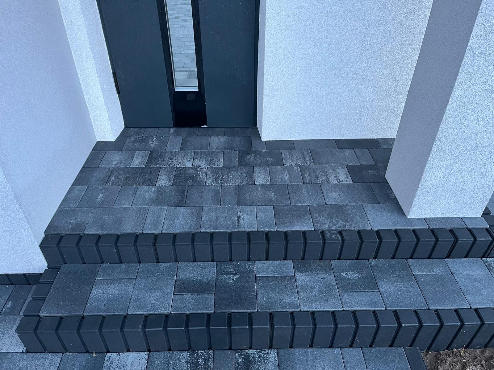 Schody wejściowe wyłożone szarą kostką brukową z ciemnymi obrzeżami, prowadzące do ciemnych drzwi wejściowych w białym budynku. Widok z góry, bliski kadr.