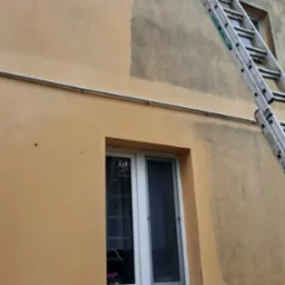 Fragment elewacji w trakcie mycia, widoczna drabina oparta o budynek, częściowo umyta ściana, okno z kwiatami na parapecie. Ujęcie z dołu.