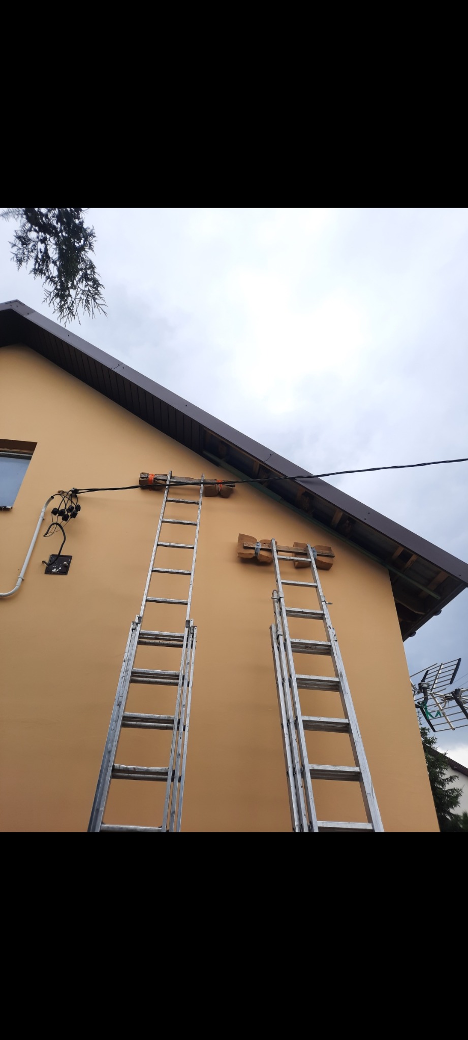 Dwie drabiny opierają się o ocieploną, pomarańczową elewację domu jednorodzinnego. Widoczne kable i mocowania. Przygotowanie do prac na wysokości.