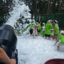 Piana party w plenerze: grupa dzieci w zielonych koszulkach bawi się w gęstej pianie wytwarzanej przez maszynę. W tle las i siatka ogrodzeniowa.