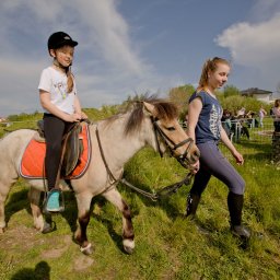 SVAGO PIOTR KUBACZYK - Dziewczynka w kasku jeździ na kucyku prowadzonym przez kobietę, w tle widoczni ludzie i ognisko na zielonym terenie. Piknik rodzinny z atrakcjami.