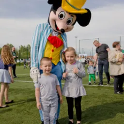 Animator w stroju Myszki Miki w żółtym kapeluszu pozuje z uśmiechniętym chłopcem i dziewczynką na zielonym boisku podczas imprezy plenerowej.