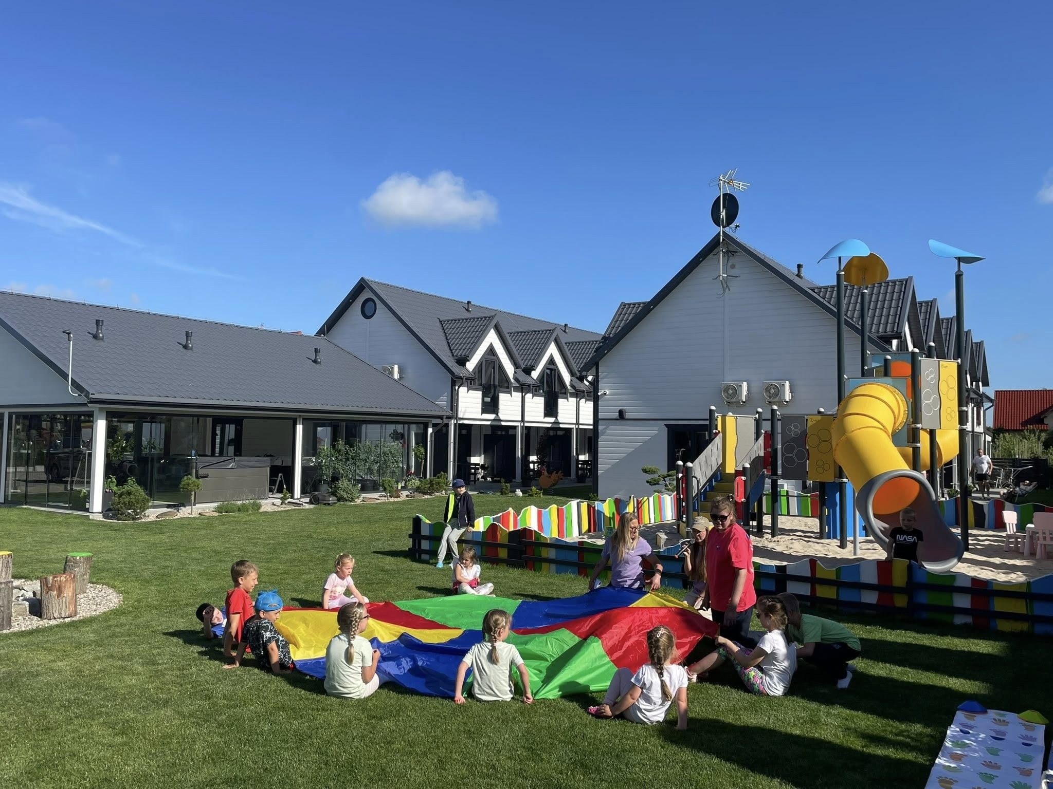 Dzieci bawią się kolorową chustą animacyjną na trawie, w tle budynki i plac zabaw. Animator w czerwonej koszulce nadzoruje zabawę. Słoneczny dzień.