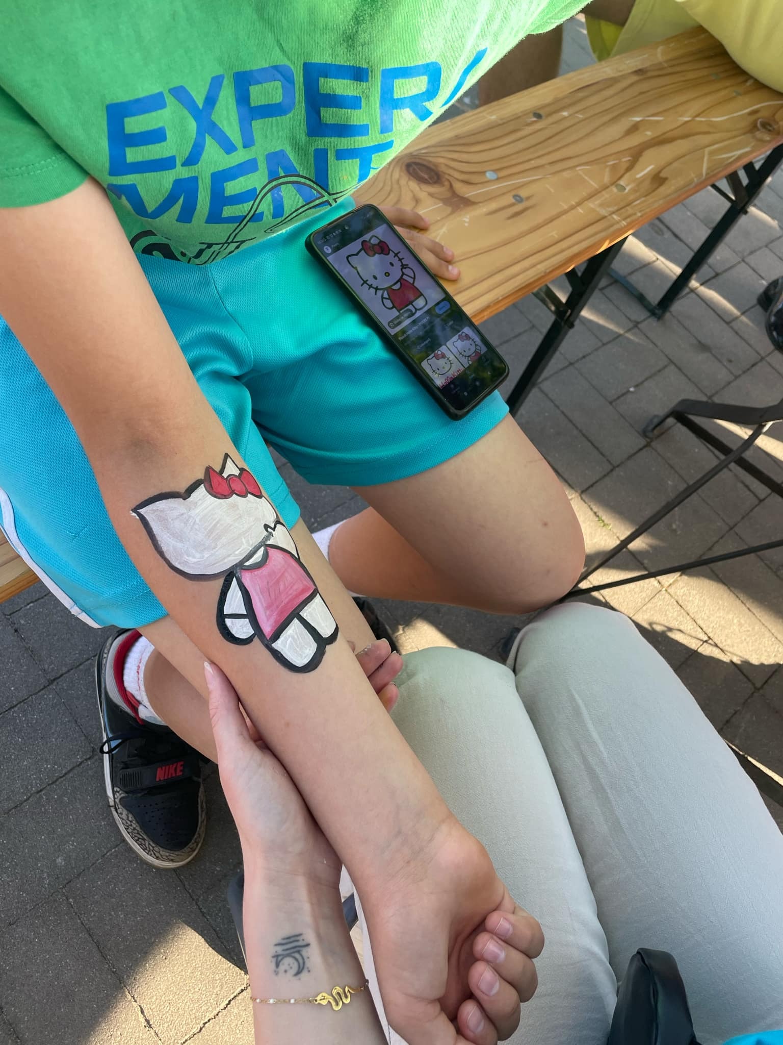 Malowanie twarzy na ramieniu dziecka, wzór Hello Kitty. Telefon z podglądem wzoru leży obok. Dziecko w zielonej koszulce i niebieskich spodenkach siedzi na drewnianej ławce.
