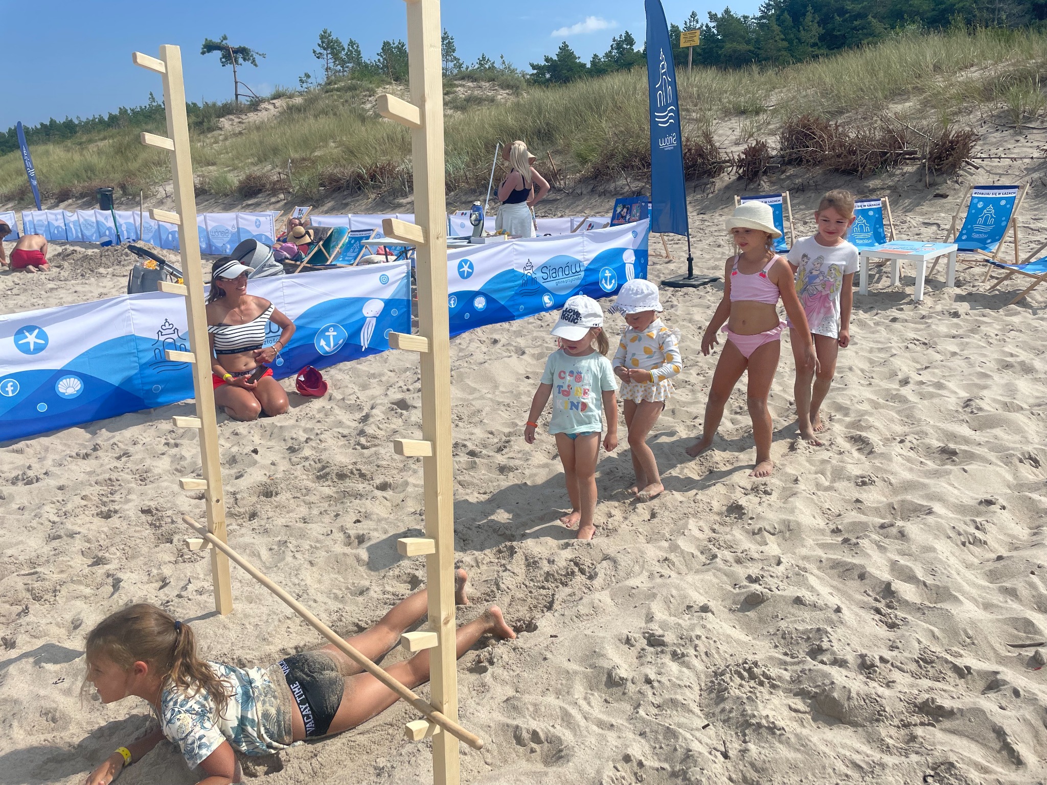 Animacje dla dzieci na plaży: dziewczynka przechodzi pod drewnianą przeszkodą, obok instruktorka i grupa dzieci. W tle parawan z logo Sianów.
