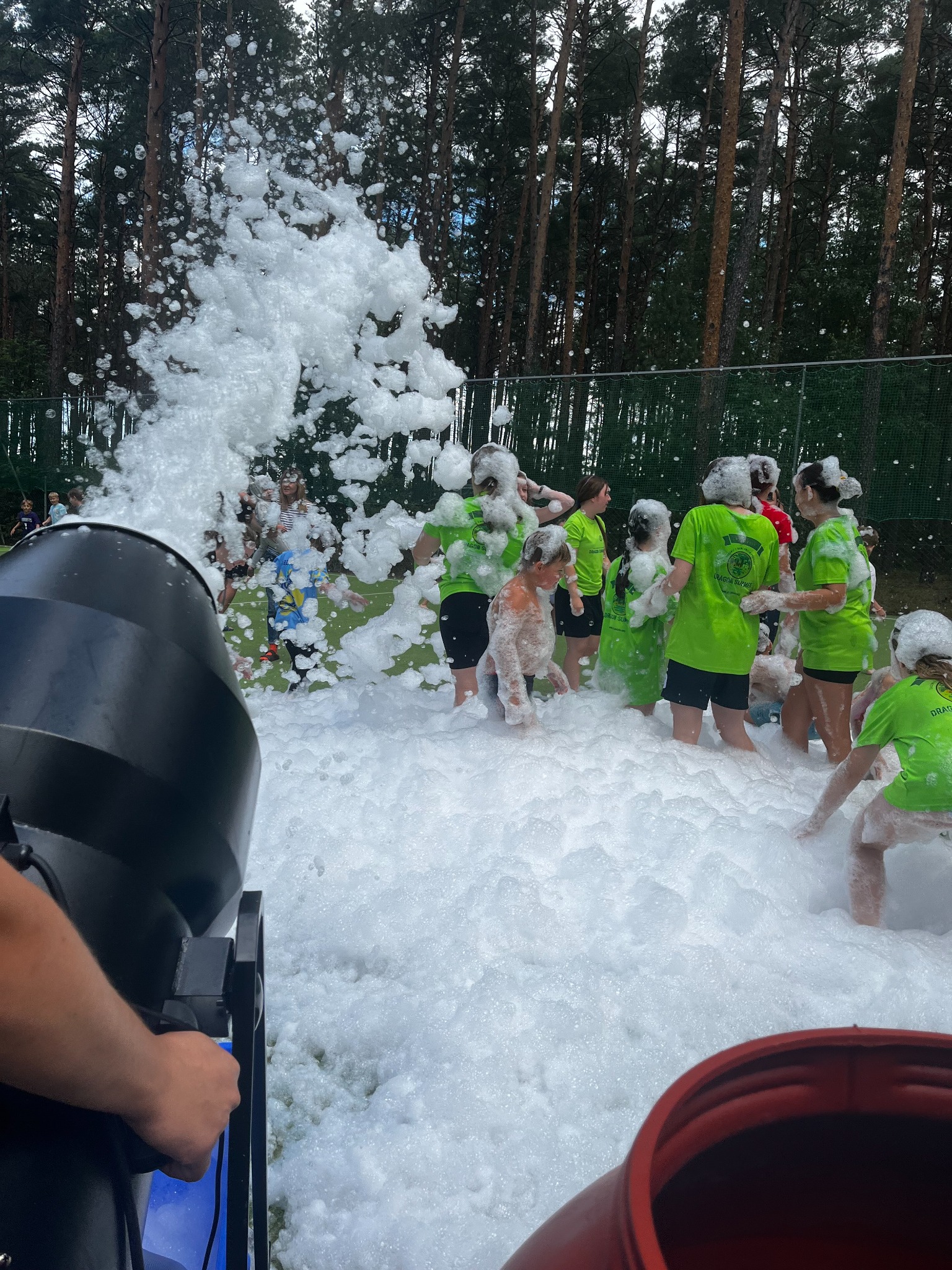 Piana party w plenerze: grupa dzieci w zielonych koszulkach bawi się w gęstej pianie wytwarzanej przez maszynę. W tle las i siatka ogrodzeniowa.