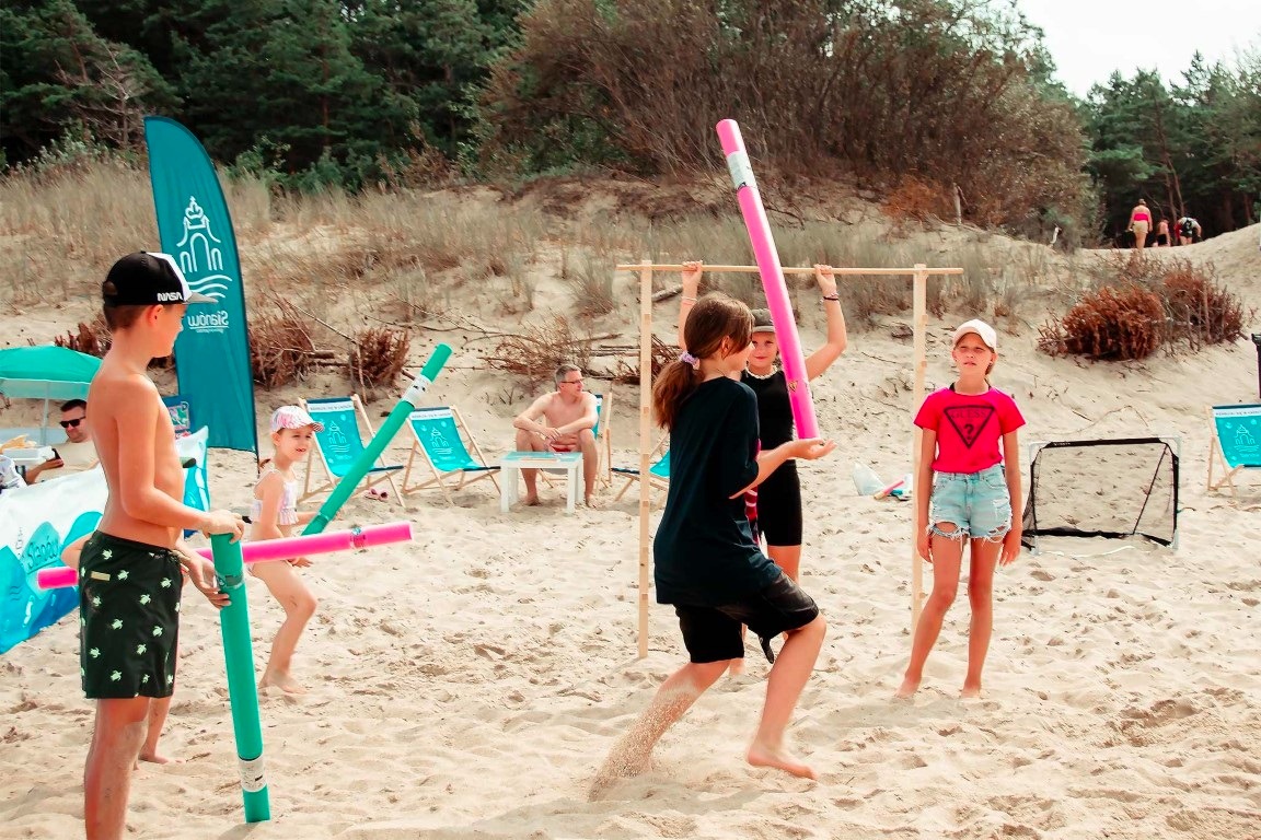 Animacja na plaży: grupa dzieci bawi się kolorowymi piankowymi pałkami pod okiem animatora. W tle leżaki, parawany i drzewa.