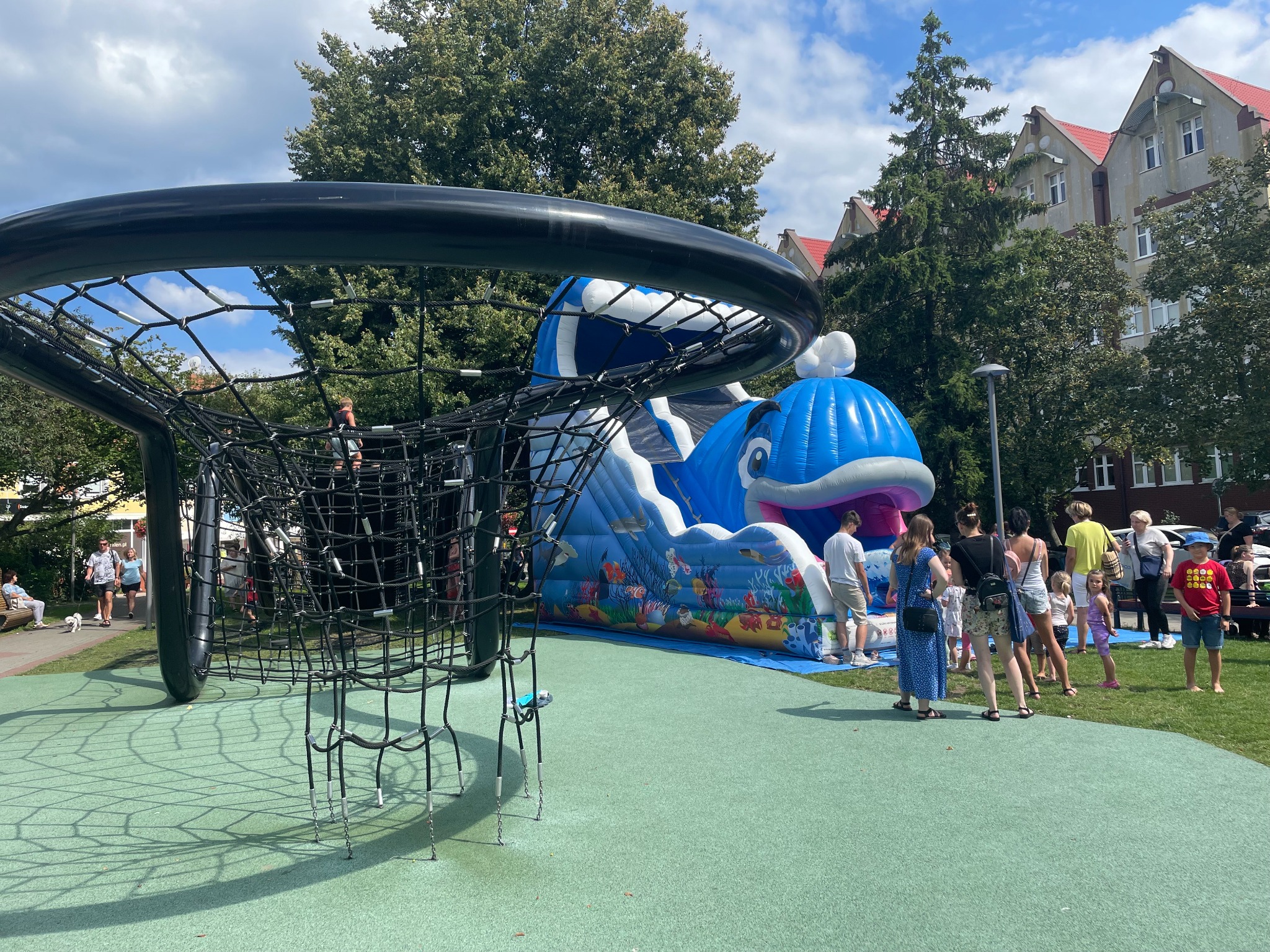 Niebieski dmuchany wieloryb na zielonej nawierzchni placu zabaw, widziany przez konstrukcję linową. W tle budynek i grupa osób.