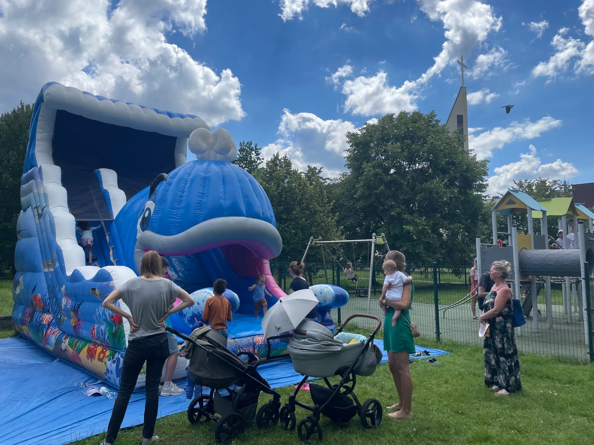 Niebieski dmuchany zamek w kształcie wieloryba na trawie, dzieci wchodzą po schodach. Rodzice obserwują. W tle kościół i plac zabaw.