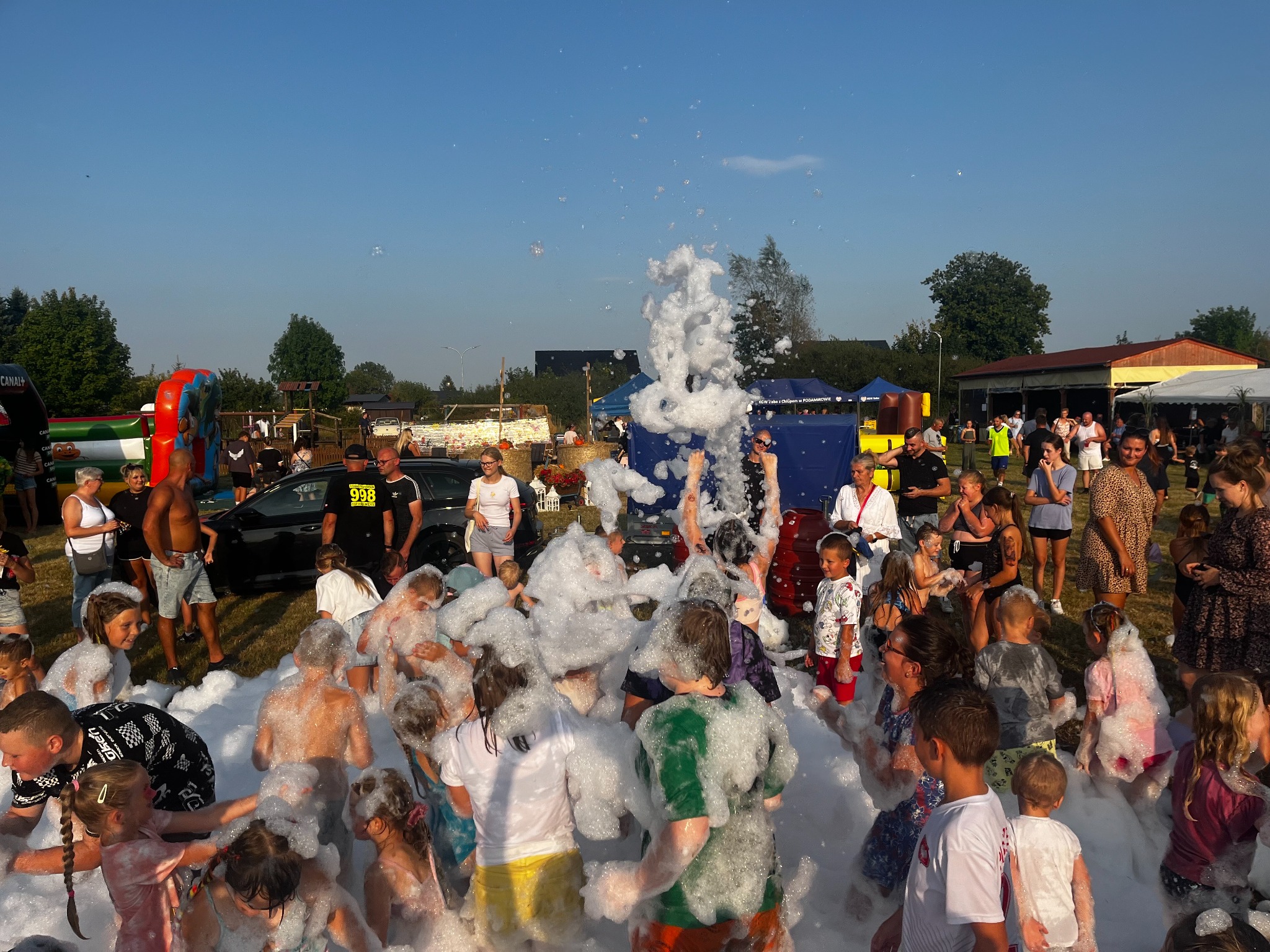 Piana party na świeżym powietrzu: grupa dzieci i dorosłych bawi się w gęstej pianie, w tle dmuchaniec i scena, słoneczny dzień.