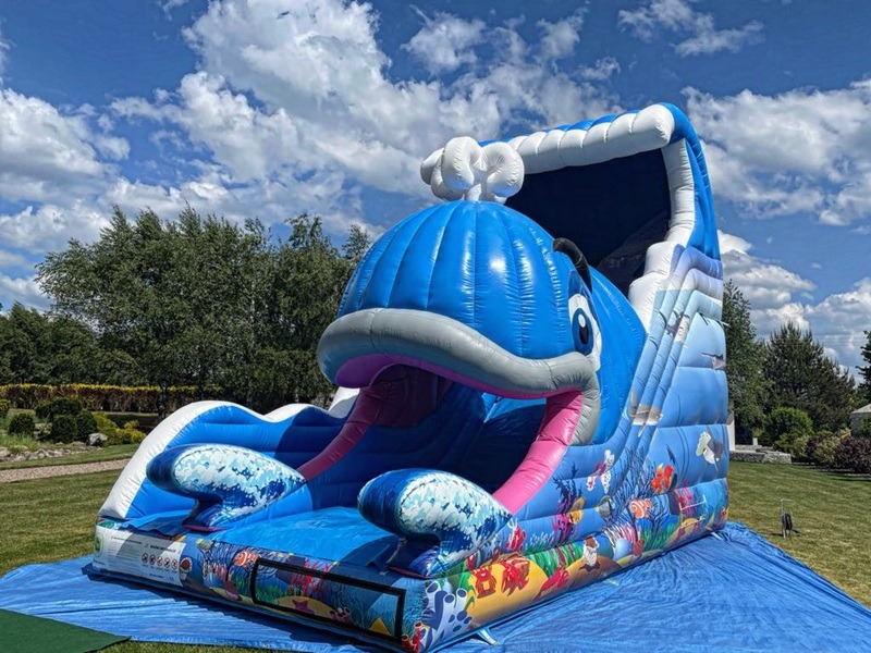 Niebieski dmuchany zamek w kształcie wieloryba na zielonej trawie, błękitne niebo w tle. Atrakcja dla dzieci z motywem morskim.