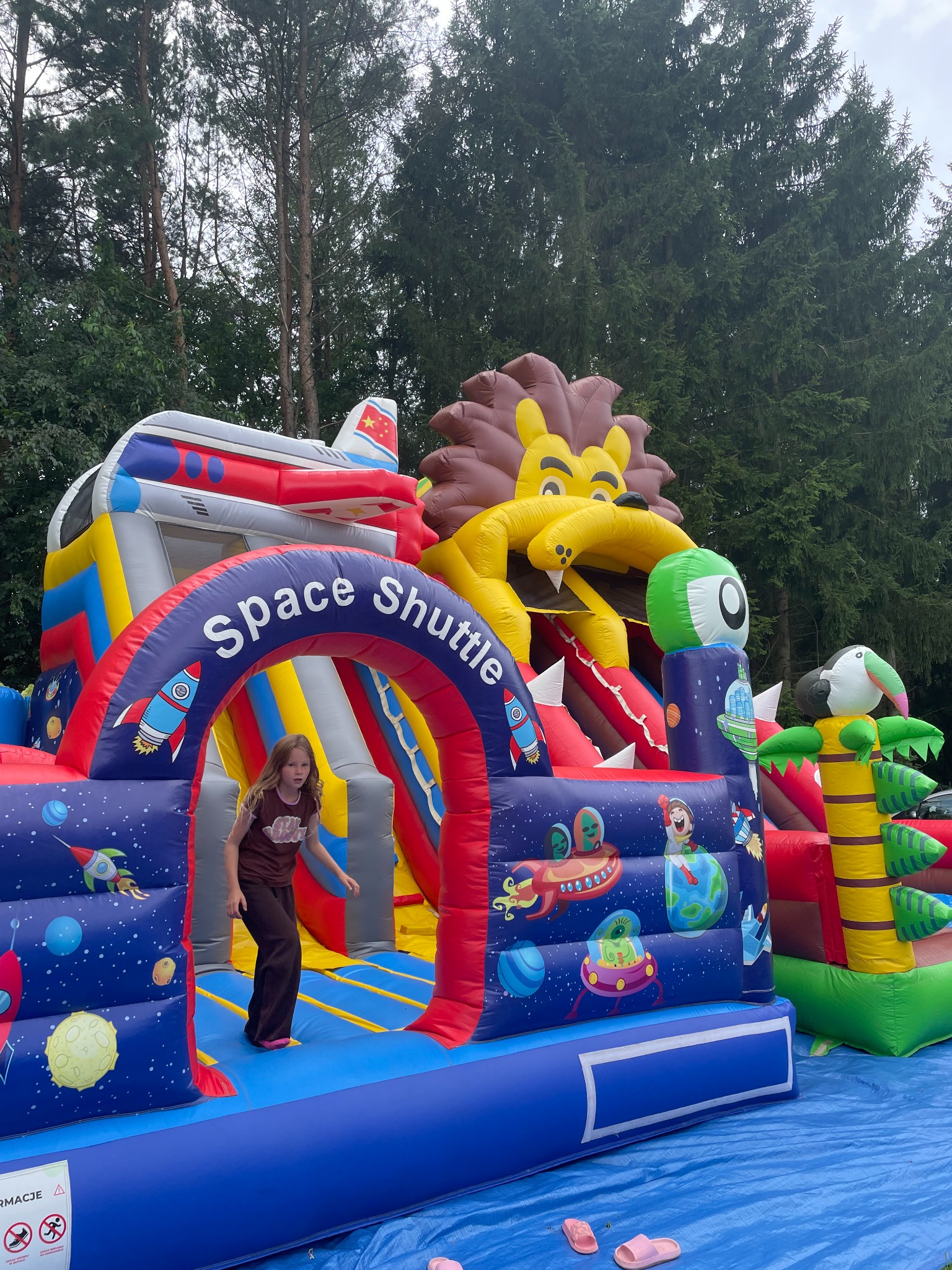 Dziewczynka wychodzi z dmuchanego zamku w kosmicznym stylu z motywem lwa, rakiet i planet, na tle drzew. Jasne kolory, atrakcja dla dzieci na świeżym powietrzu.