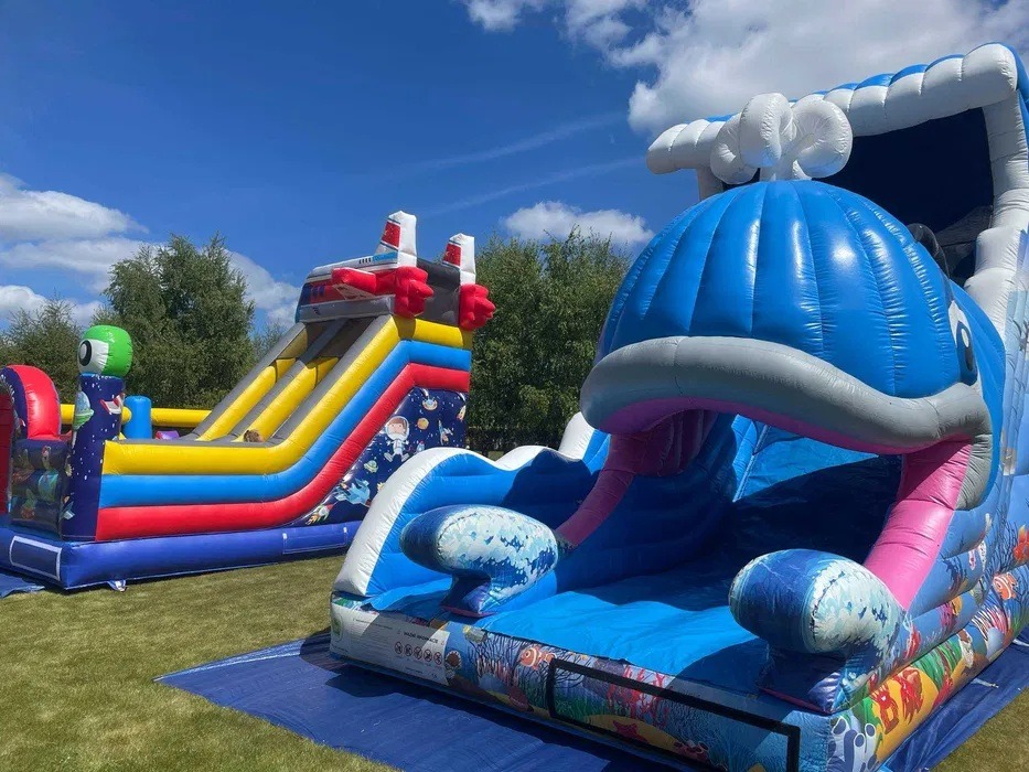 Dwa dmuchańce: statek kosmiczny z motywem kosmosu i zjeżdżalnia w kształcie niebieskiego wieloryba na trawie w słoneczny dzień.