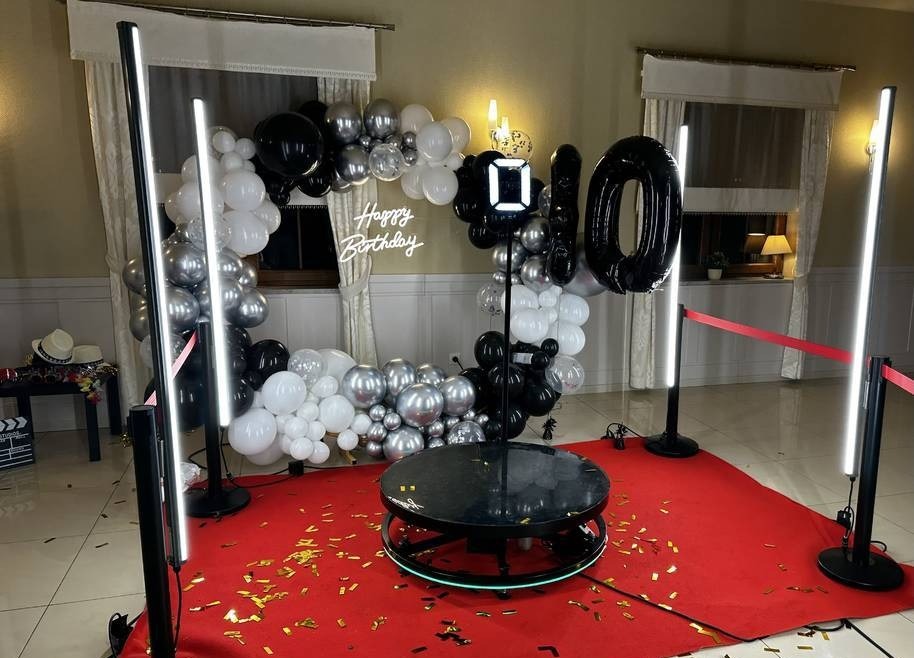 Kompletna instalacja fotobudki 360 z dekoracjami balonowymi (czarno-srebrne balony, napis 'Happy Birthday') na czerwonym dywanie z konfetti.