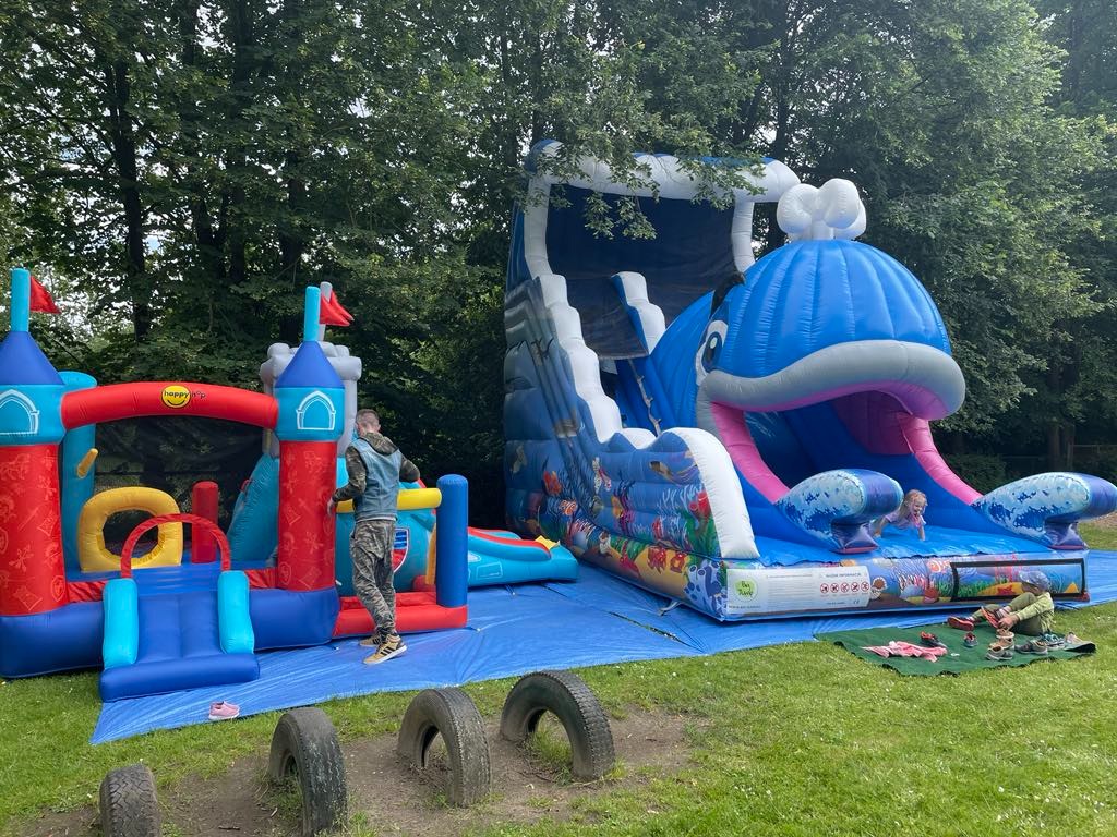 Dwa dmuchańce: zamek z tunelem i zjeżdżalnia w kształcie wieloryba, rozłożone na trawie. Dzieci bawią się na dmuchańcach i obok nich. Opony jako element dekoracyjny.