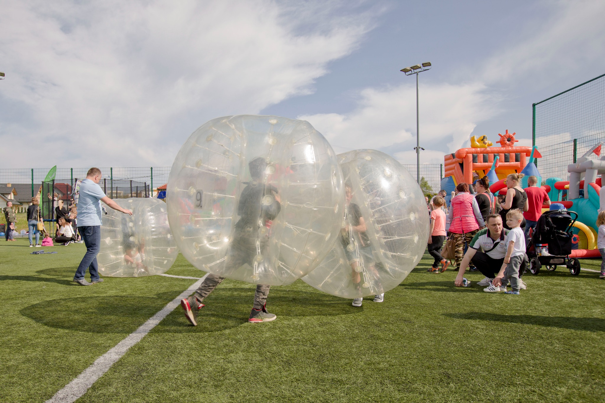 Uczestnicy w przezroczystych kulach na zielonej trawie, w tle dmuchany plac zabaw i widzowie. Zabawa na świeżym powietrzu podczas imprezy plenerowej.