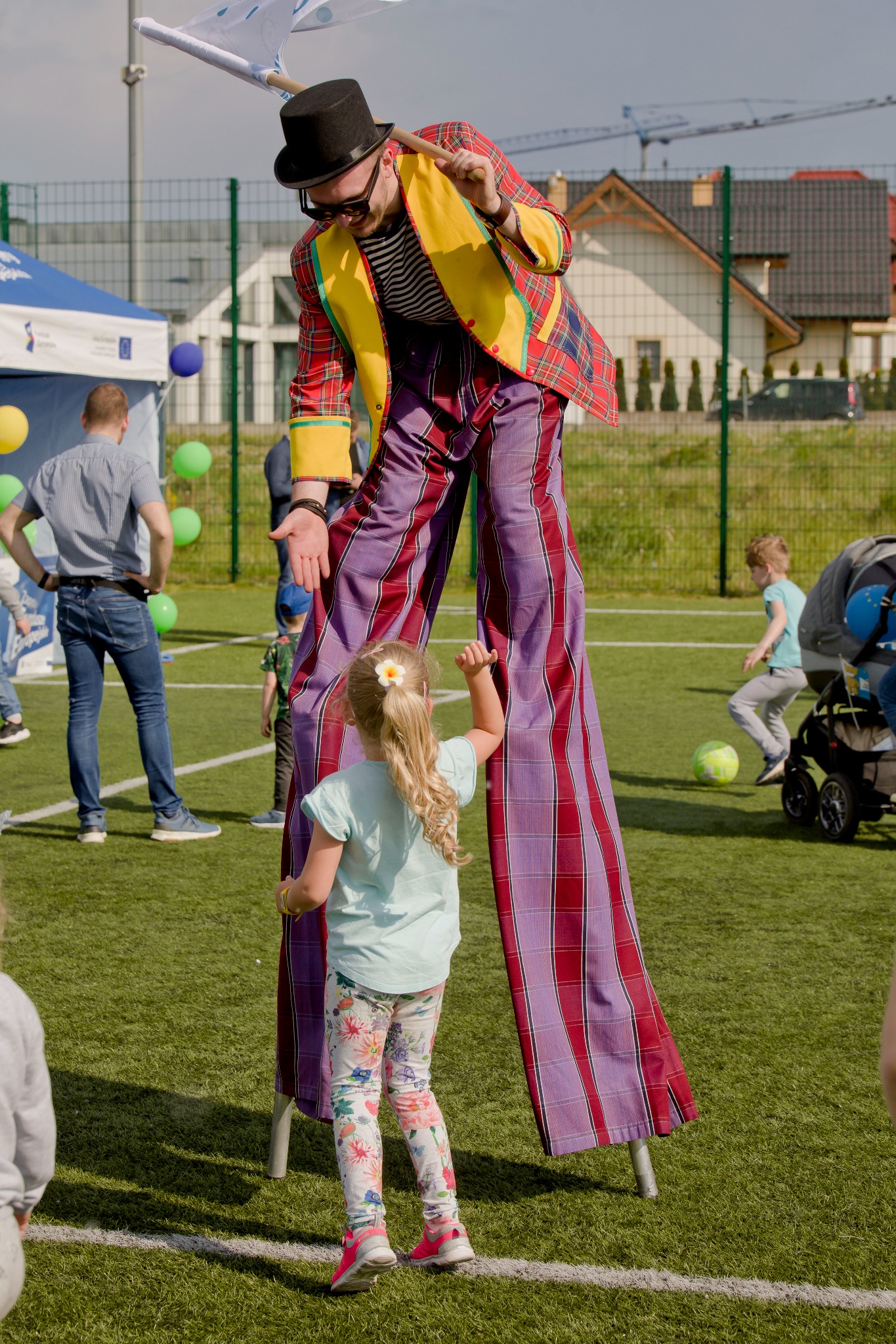 Animator na szczudłach w kolorowym stroju, interakcja z dzieckiem na tle zielonego trawnika. Widoczne elementy imprezy plenerowej, domy w tle i ogrodzenie.