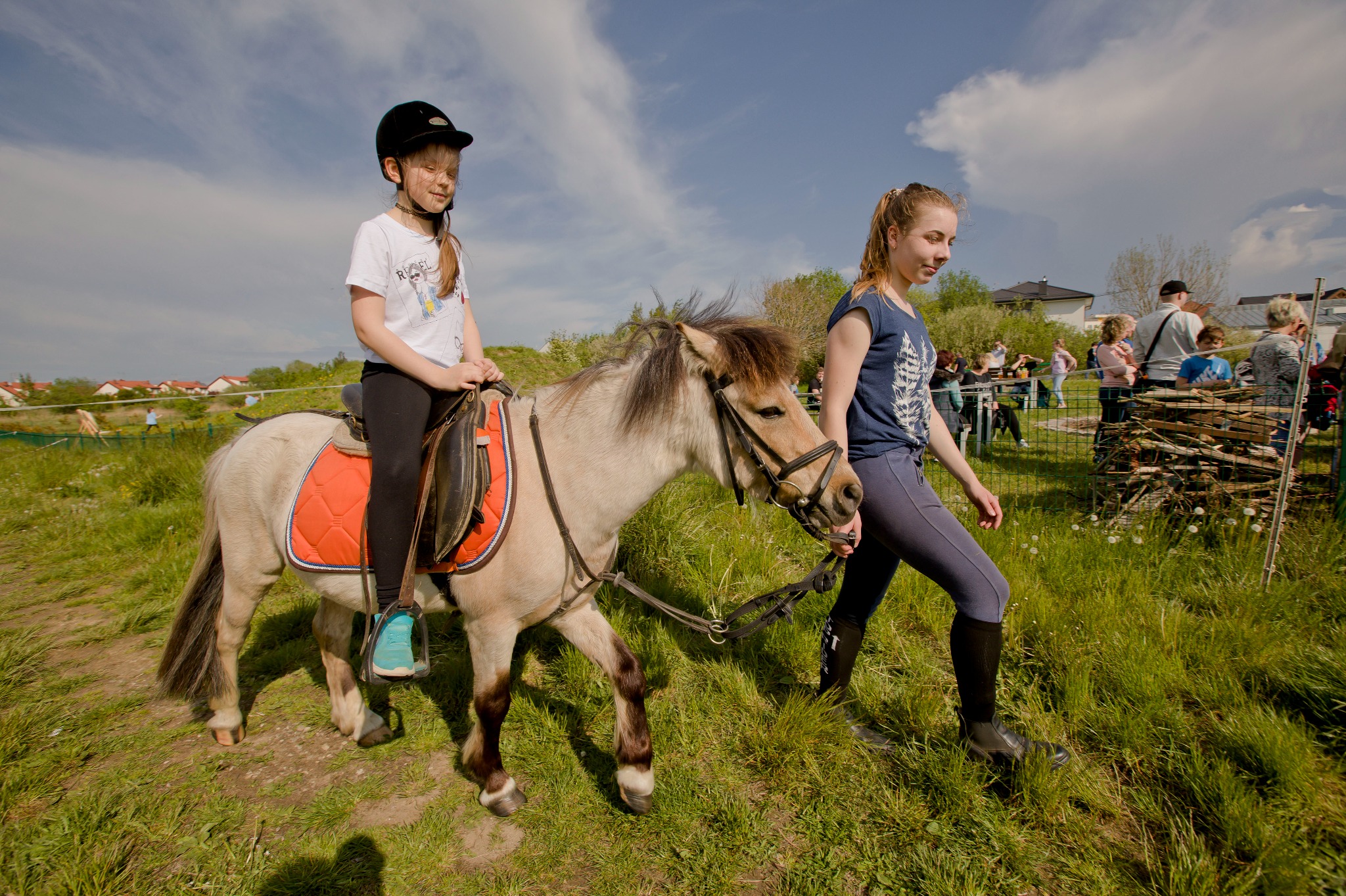Dziewczynka w kasku jeździ na kucyku prowadzonym przez kobietę, w tle widoczni ludzie i ognisko na zielonym terenie. Piknik rodzinny z atrakcjami.