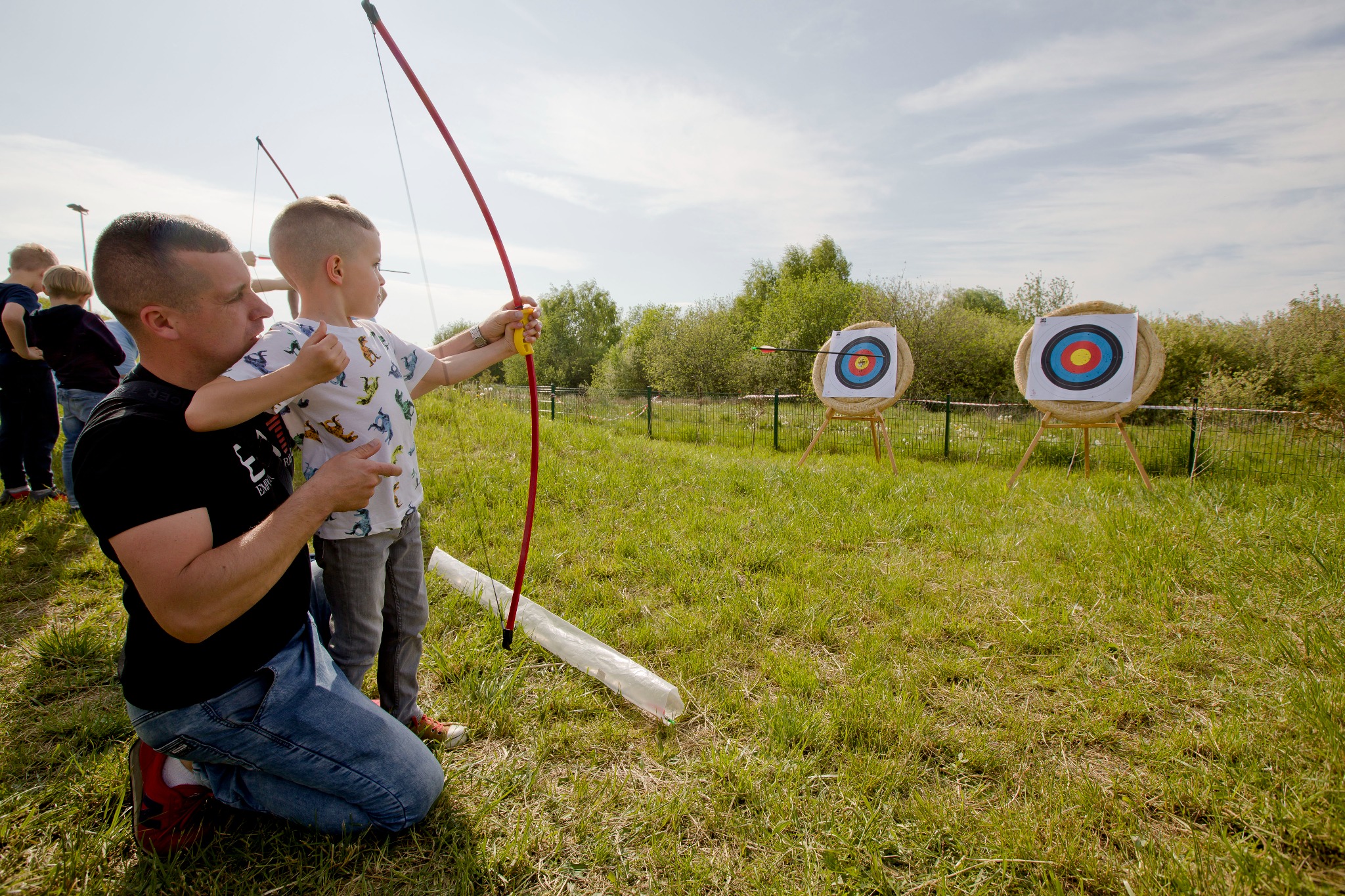 Instruktor uczy chłopca strzelać z łuku na tle trawnika z dwoma tarczami. Jedna strzała trafiła w cel. Dzieci obserwują z boku. Słoneczny dzień.