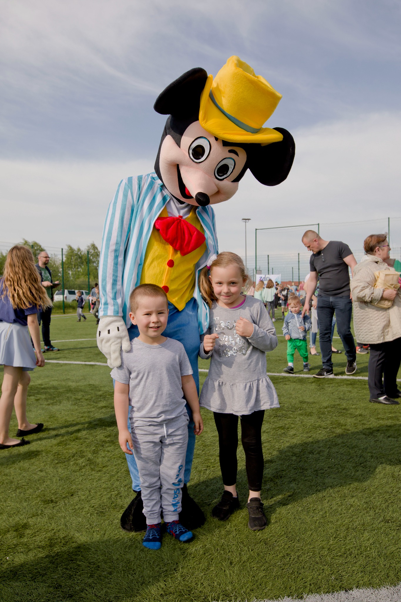 Animator w stroju Myszki Miki w żółtym kapeluszu pozuje z uśmiechniętym chłopcem i dziewczynką na zielonym boisku podczas imprezy plenerowej.