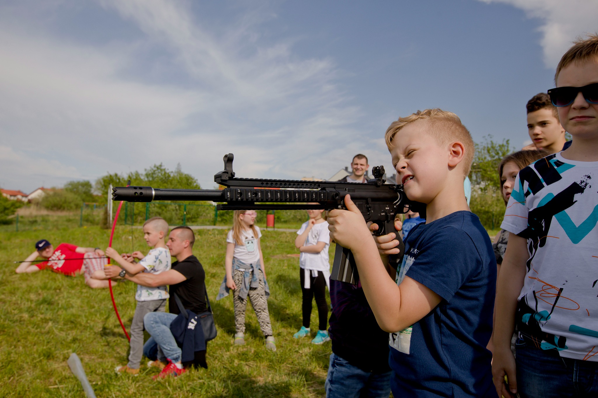 Grupa dzieci, chłopiec celuje z repliki karabinu na tle zielonej trawy i nieba, inni uczestniczą w zabawie z łukami, aktywne spędzanie czasu na świeżym powietrzu.