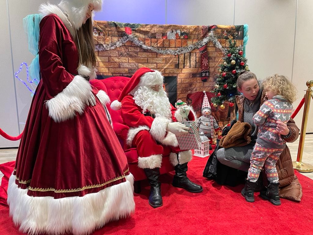 Spotkanie dziecka z Mikołajem w świątecznej scenerii z choinką i postacią w stroju diablicy. Czerwony dywan i fotel tworzą ciepły klimat.