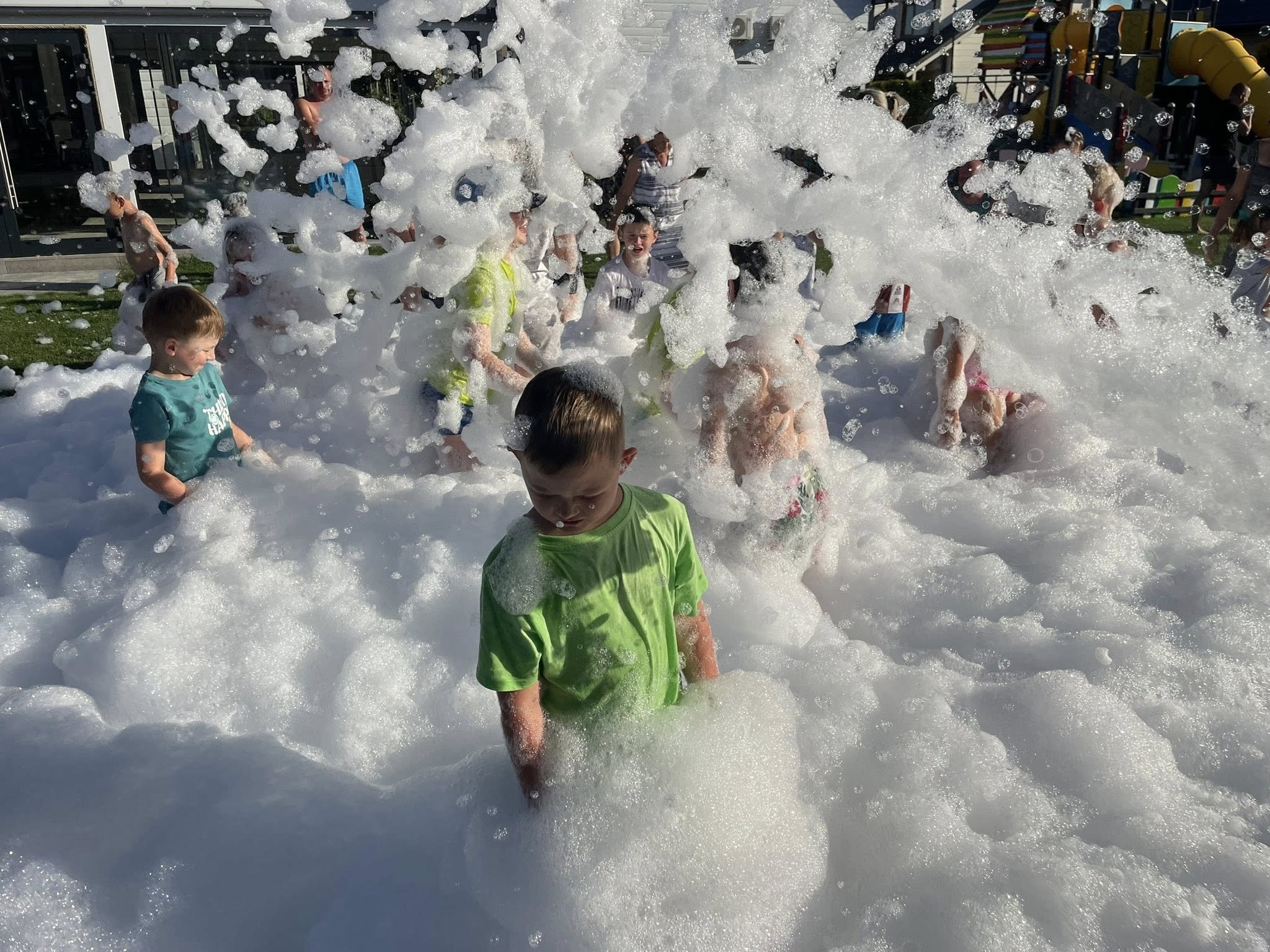 Dzieci bawiące się w pianie na zewnątrz, z placem zabaw w tle. Impreza plenerowa z użyciem wytwornicy piany, tworzącej efekt śnieżnej zabawy w letni dzień.