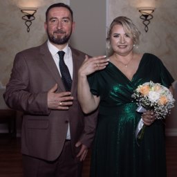 Hamad photography - Uśmiechnięta para w eleganckich strojach: mężczyzna w brązowym garniturze, kobieta w szmaragdowej sukni trzyma bukiet ślubny. Widoczne obrączki.