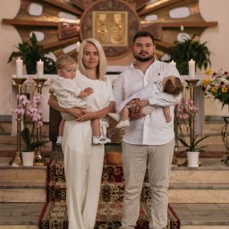 Hamad photography - Portret rodzinny w kościele: rodzice w bieli trzymają dwoje małych dzieci na tle ołtarza i kwiatów. Uwagę zwracają białe ubrania i obuwie sportowe.