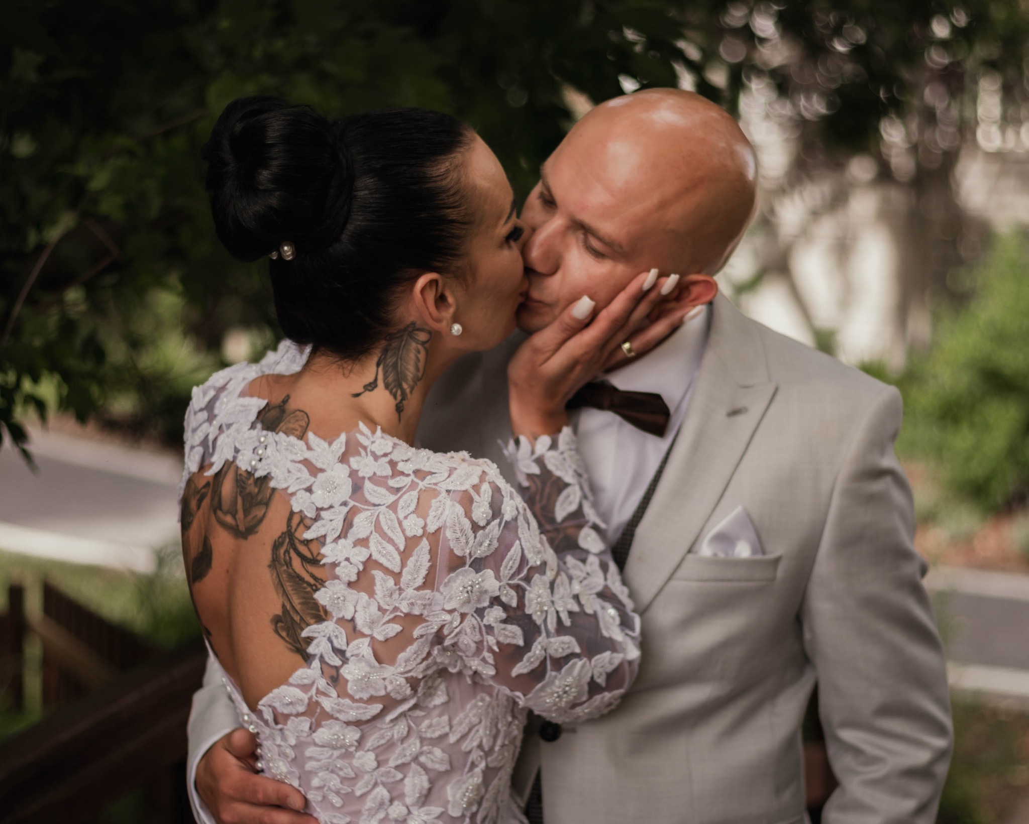 Pocałunek nowożeńców: panna młoda w koronkowej sukni z tatuażami na plecach i szyi, pan młody w szarym garniturze z muchą, w tle zieleń.