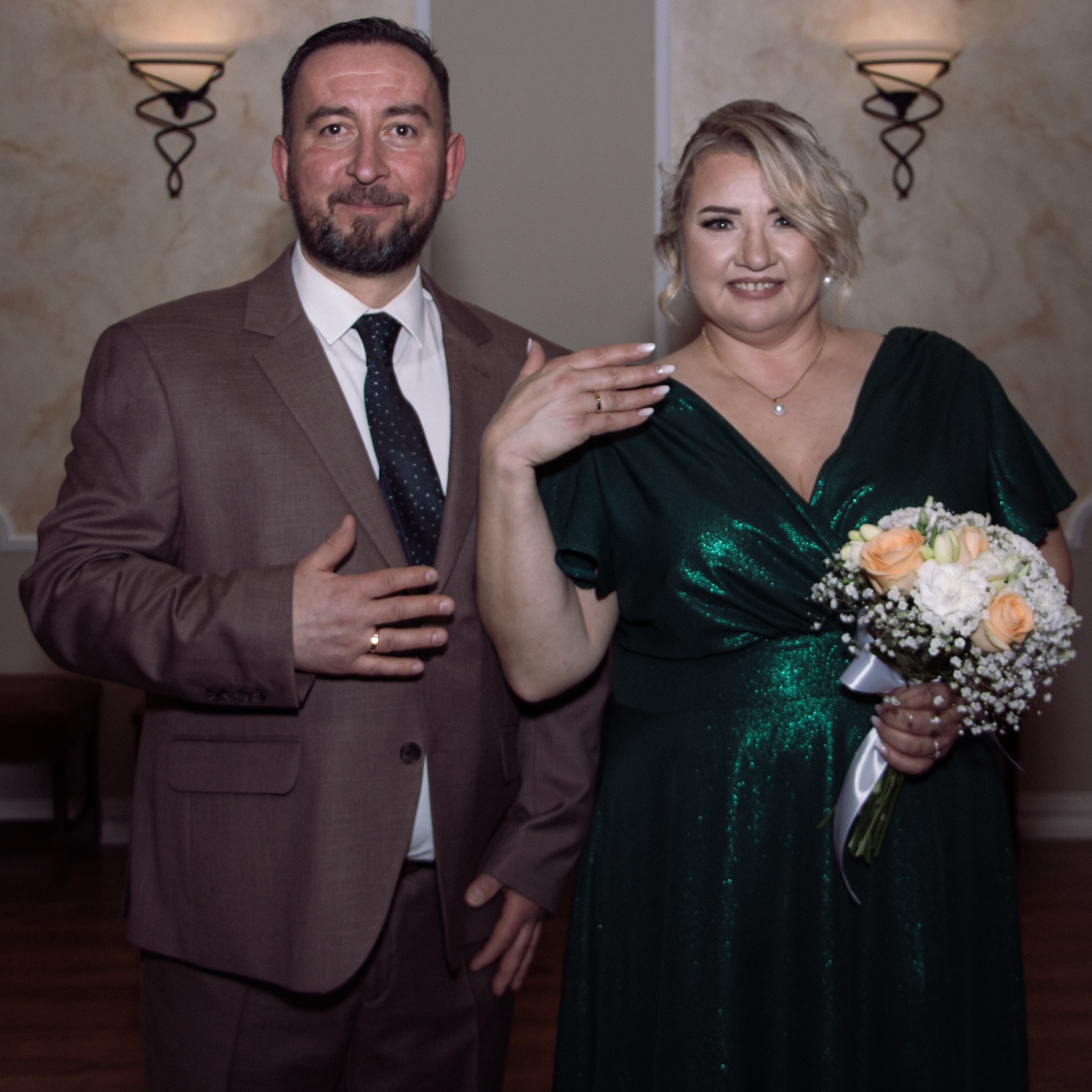 Uśmiechnięta para w eleganckich strojach: mężczyzna w brązowym garniturze, kobieta w zielonej sukni trzyma bukiet. Widoczne obrączki ślubne, dekoracyjne oświetlenie w tle.