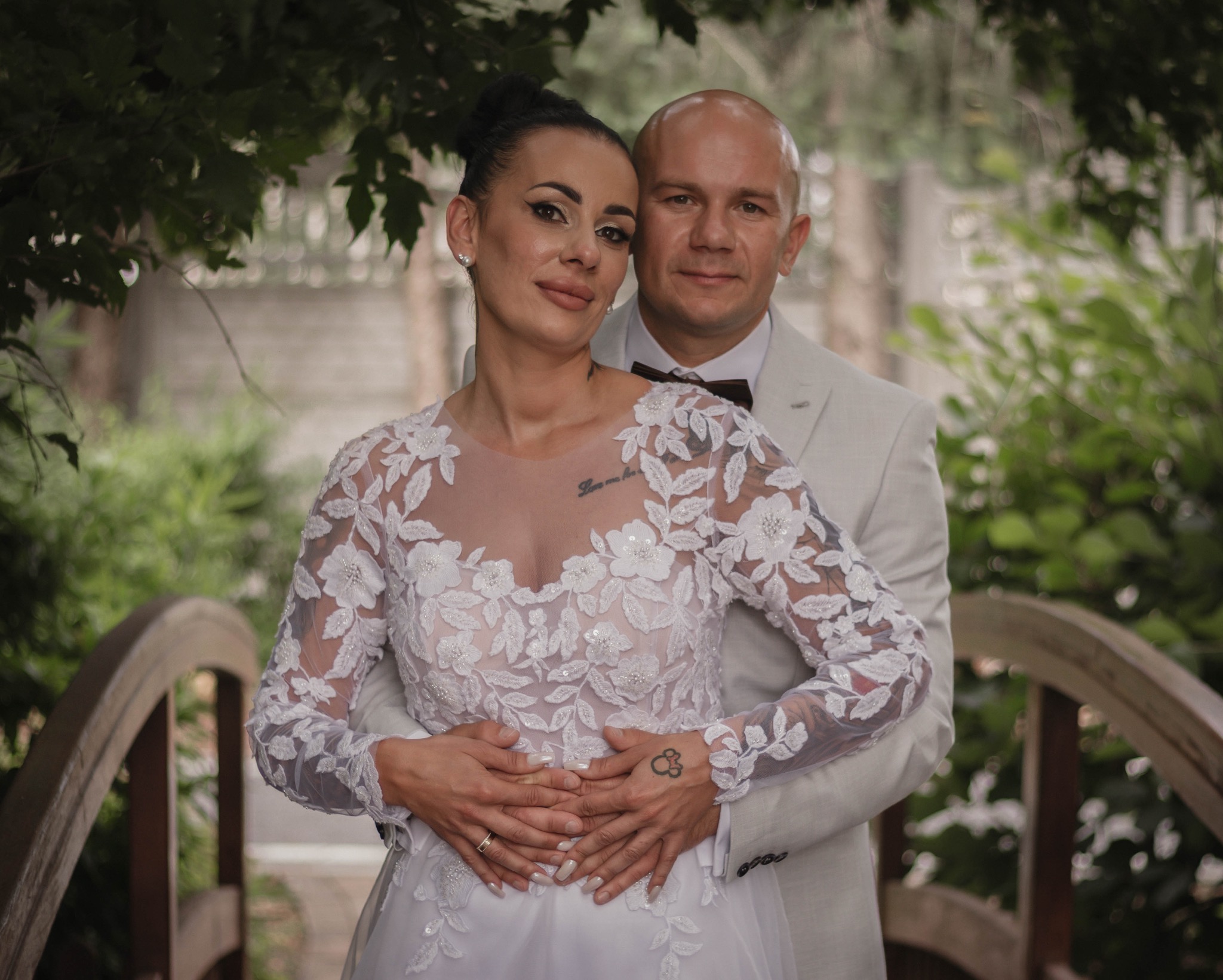 Para młoda w objęciach na drewnianym mostku w plenerze. Panna młoda w sukni z koronkowymi aplikacjami, pan młody w jasnym garniturze z muchą. Romantyczna atmosfera.