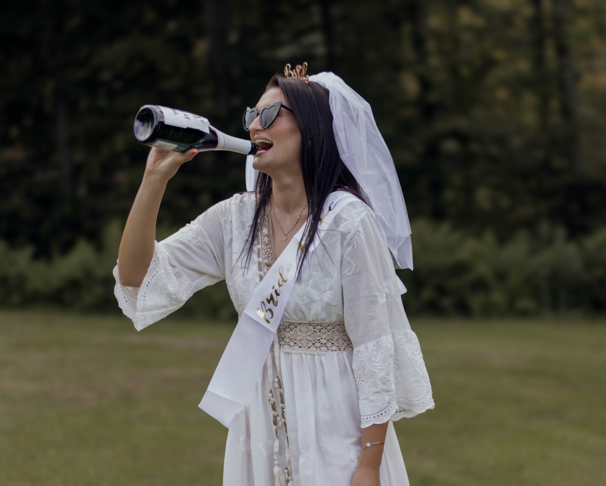 Panna młoda w okularach w kształcie serc pije szampana z butelki, ubrana w białą sukienkę i welon na tle zieleni.