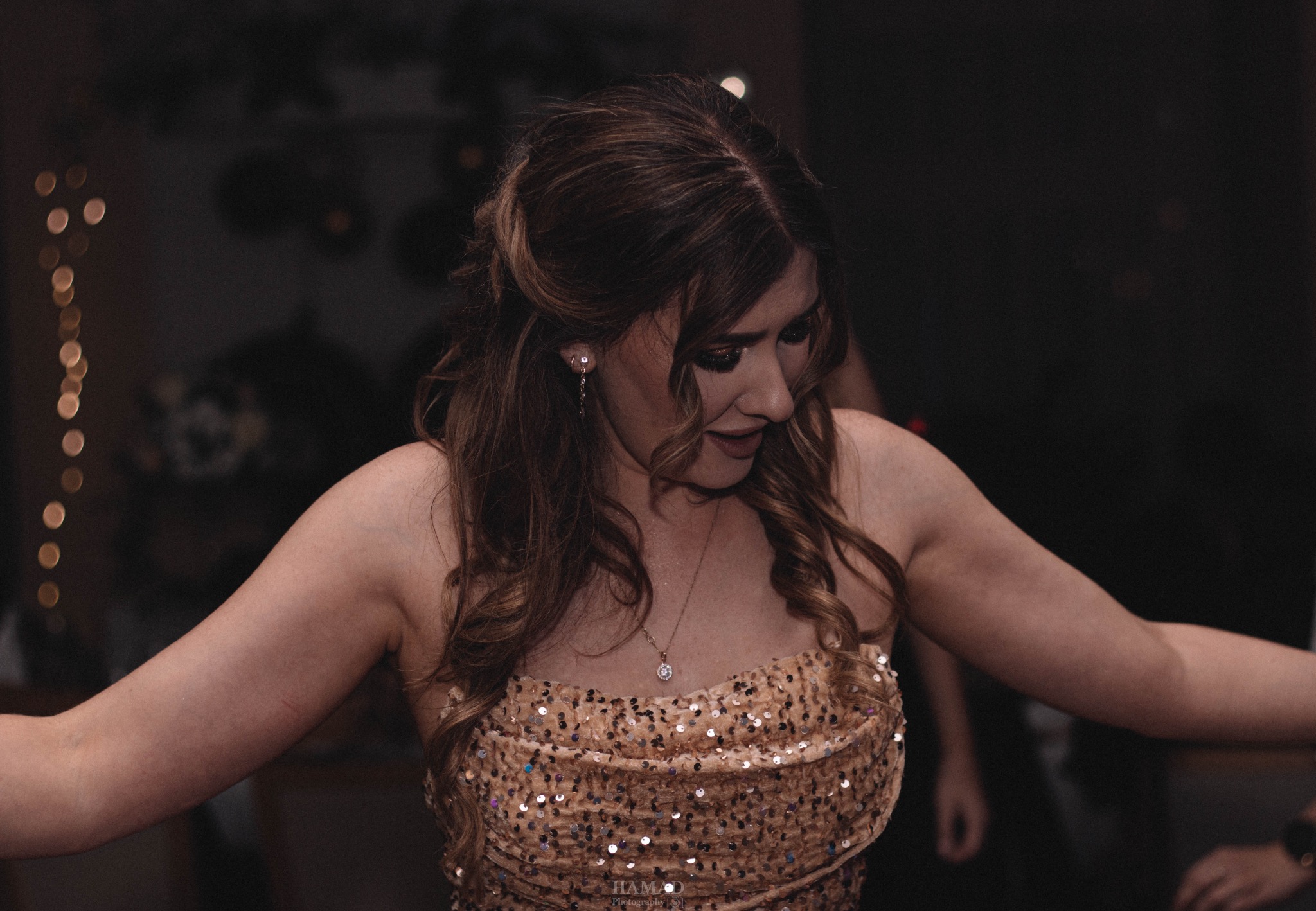 Kobieta w cekinowej sukience, z upiętymi włosami i delikatną biżuterią, spogląda w dół. W tle ciemne otoczenie z rozproszonymi światłami. Ujęcie z bliska.