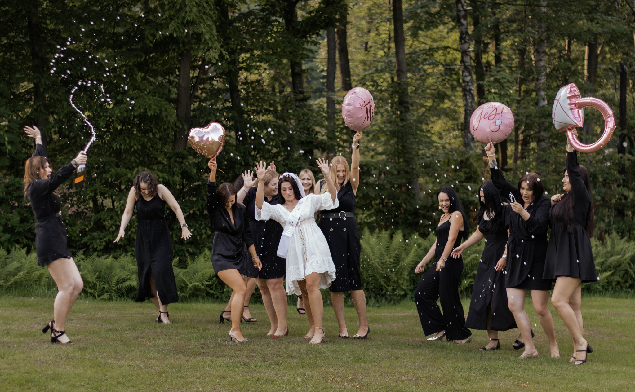 Wieczór panieński w plenerze: przyszła panna młoda w bieli, druhny w czerni, konfetti i balony w kształcie serca i pierścionka.