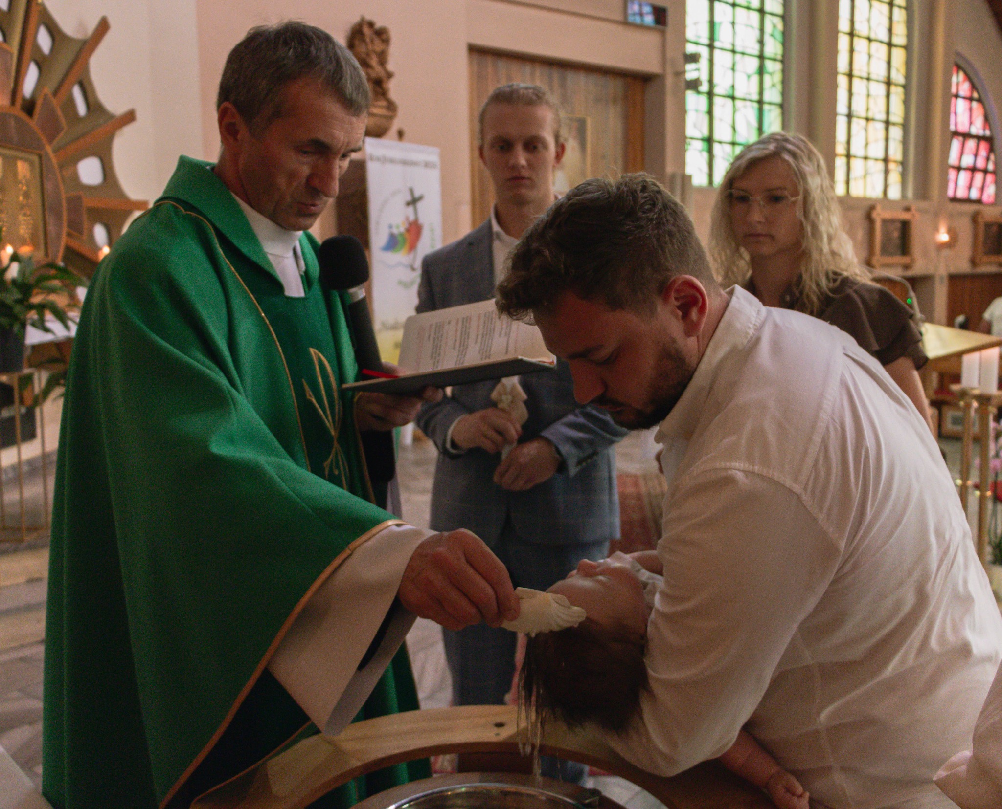 Ksiądz w zielonej szacie udziela sakramentu chrztu dziecku w kościele. W tle rodzice i ministrant.
