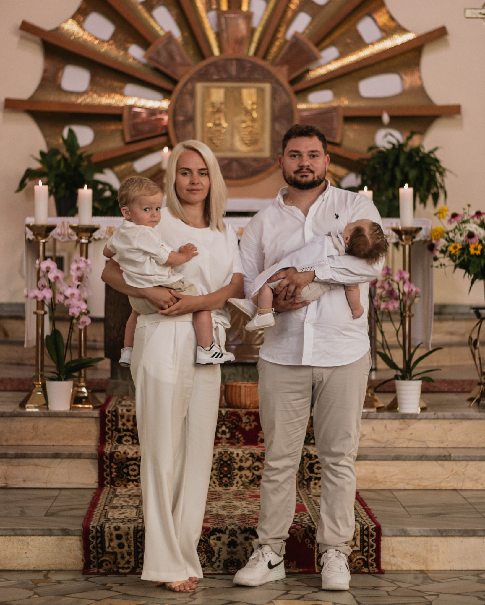 Portret rodzinny w kościele: rodzice w bieli trzymają dwoje małych dzieci na tle ołtarza i kwiatów. Uwagę zwracają białe ubrania i obuwie sportowe.
