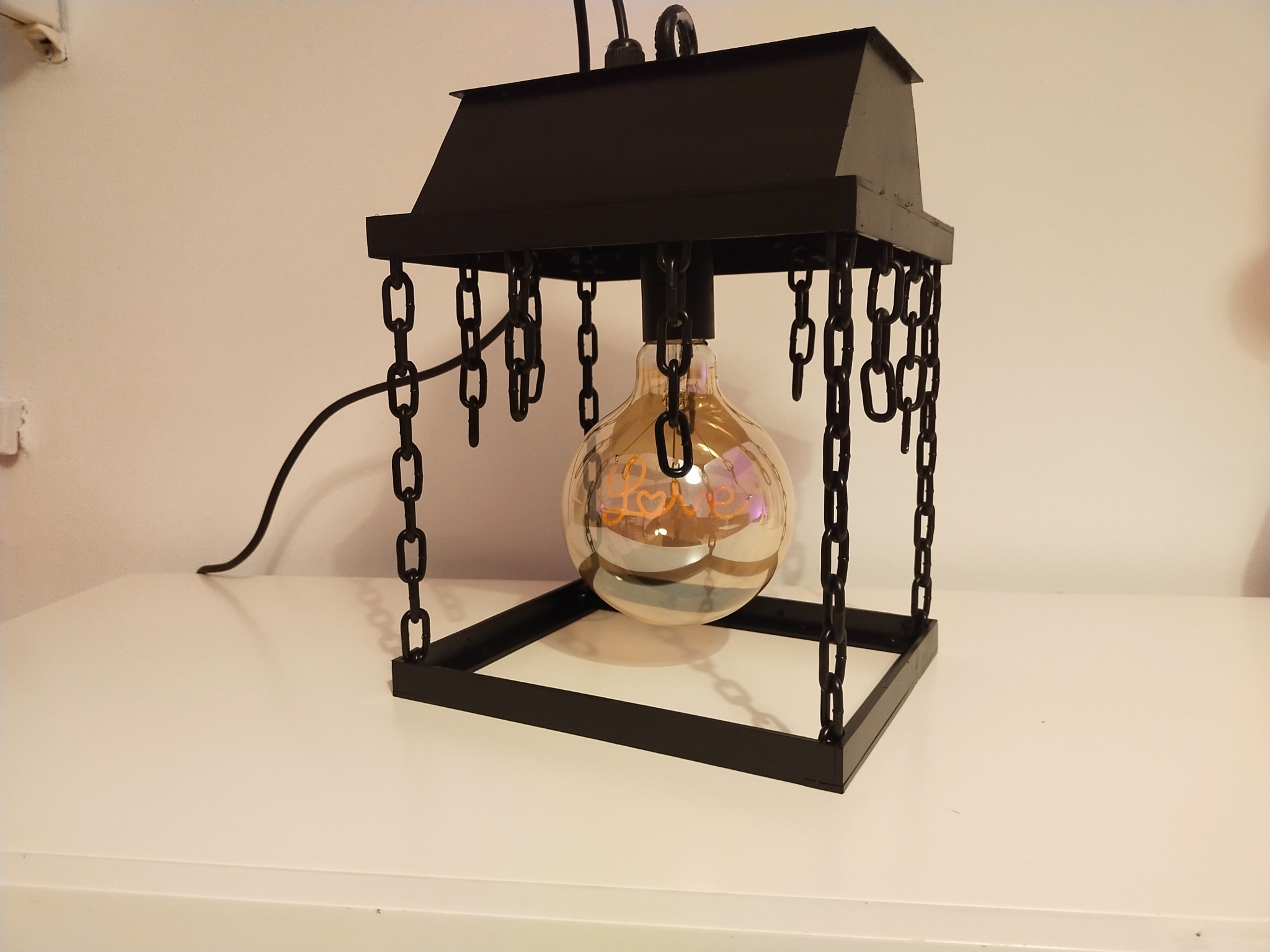 Czarna, metalowa lampa wisząca w stylu industrialnym z łańcuchami i żarówką dekoracyjną z napisem LOVE, stojąca na białym blacie.