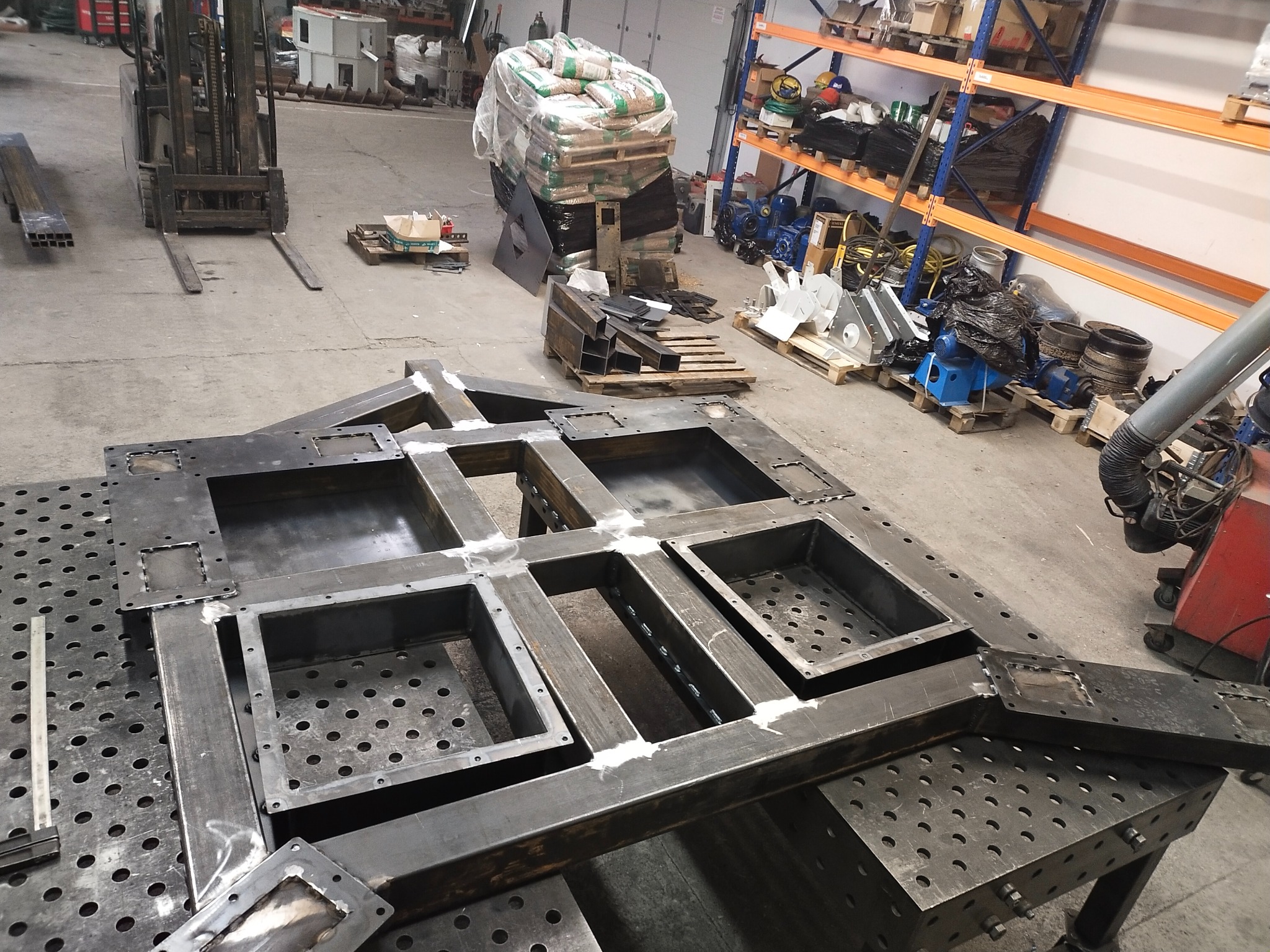 Konstrukcja metalowa z profili spawanych na stole spawalniczym. Widoczny wózek widłowy, regał z narzędziami i materiałami. Hala produkcyjna.