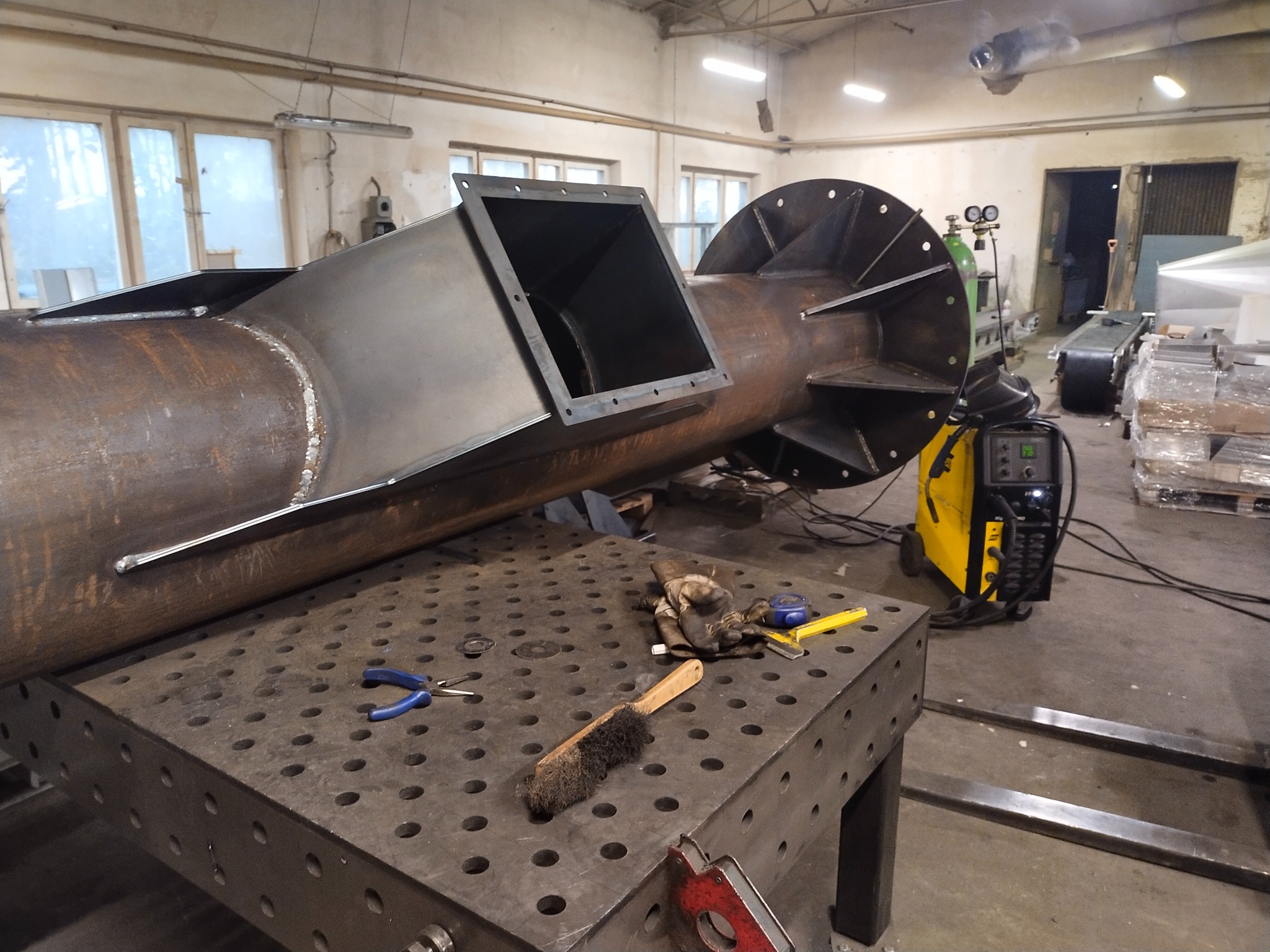 Metalowy komin w trakcie budowy w warsztacie. Widoczny stół spawalniczy, narzędzia i spawarka. Duży element konstrukcyjny z blachy.