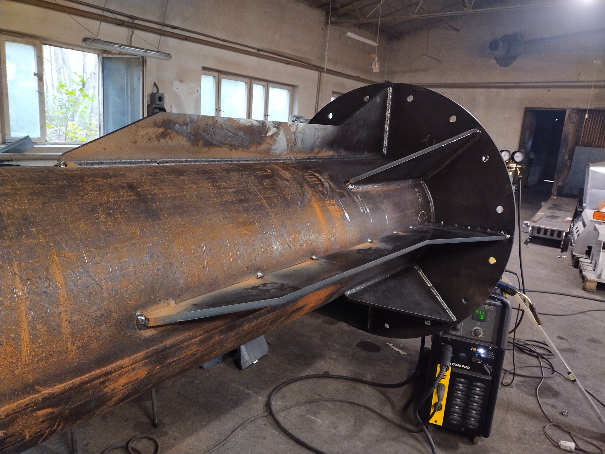 Metalowa konstrukcja komina w trakcie spawania, widoczne spoiny i elementy mocujące, surowy industrialny wygląd. W tle okno i sprzęt spawalniczy.