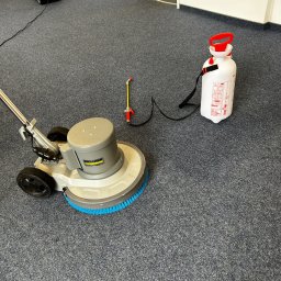 Don Clean - Maszyna Karcher z niebieską szczotką i spryskiwacz na szarym dywanie w trakcie czyszczenia. Widoczny odkurzacz. W tle białe ściany i otwarte drzwi.